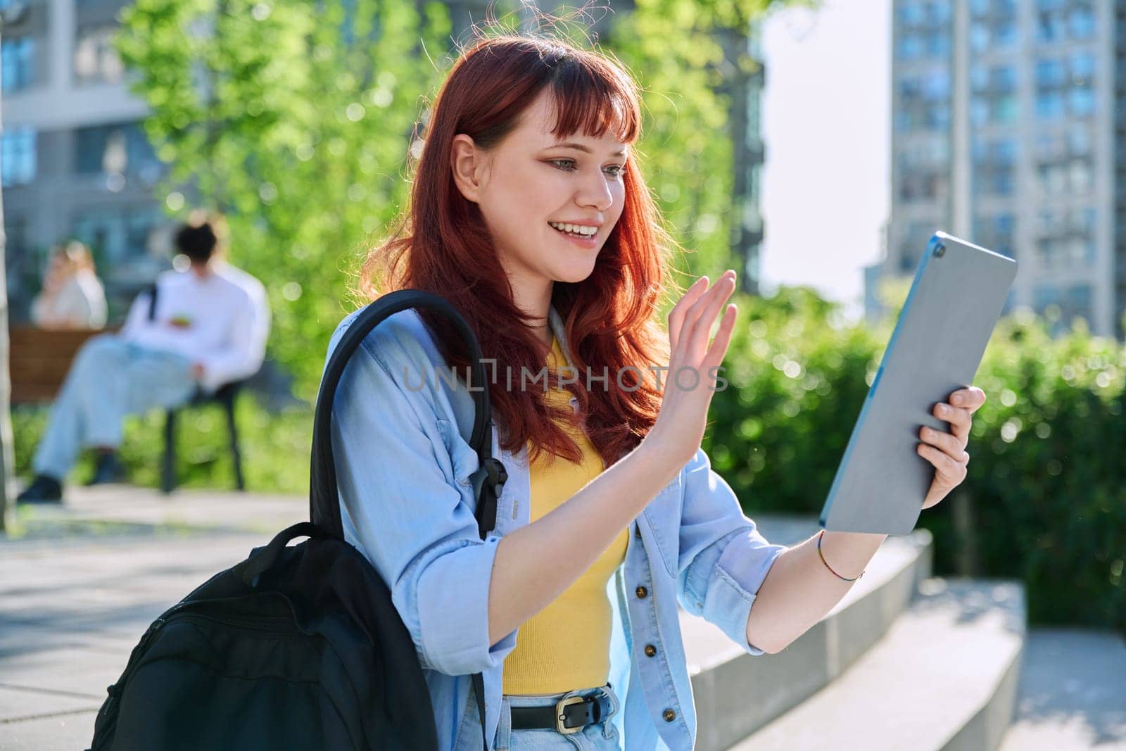 Young attractive female college student using digital tablet outdoor, talking on video call chat conference, educational building background. Education, technology, training, 19,20 years age youth concept