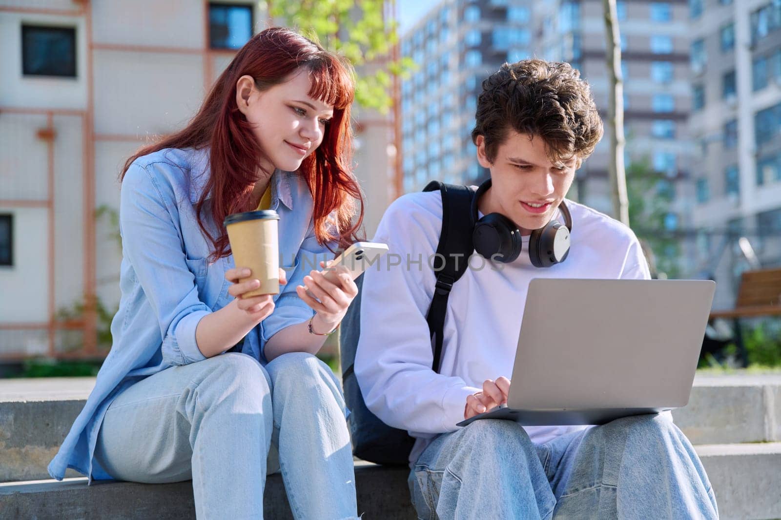 Teenage college students guy and girl talking, using laptop smartphone, sitting outdoor near educational building. Youth 19-20 years old, education, technologies, lifestyle, friendship concept