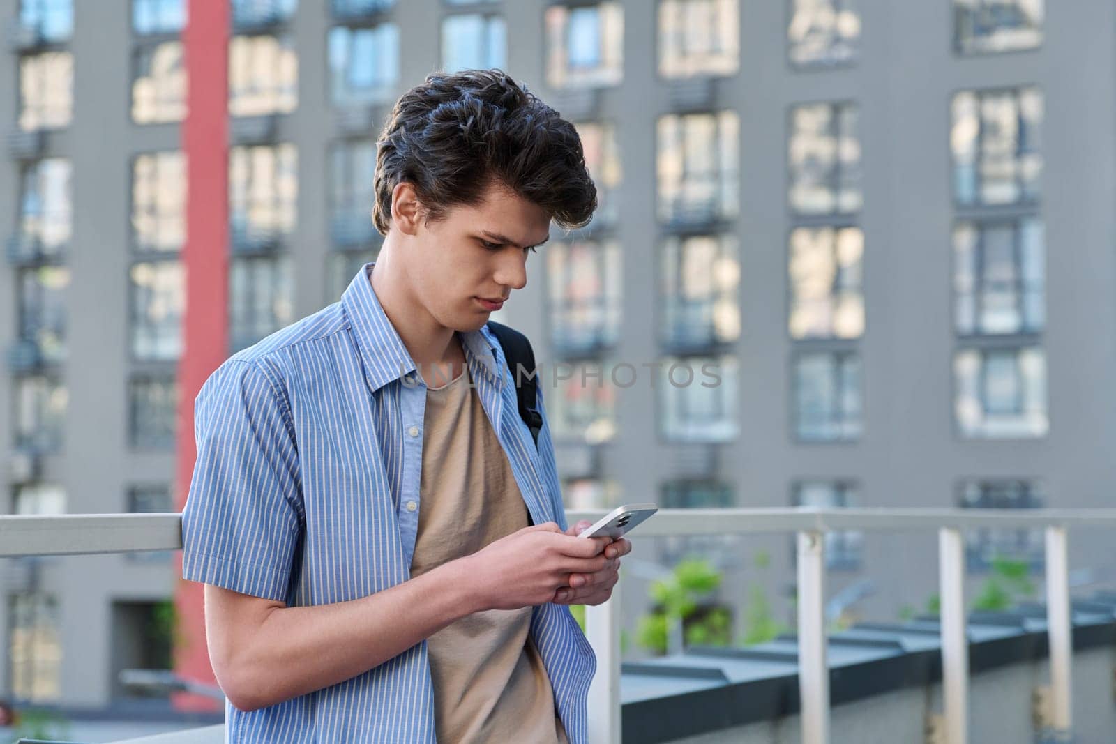Relaxed resting handsome young male with smartphone, urban outdoor, modern building background. Student 19-20 years old using phone, texting, lifestyle, youth concept
