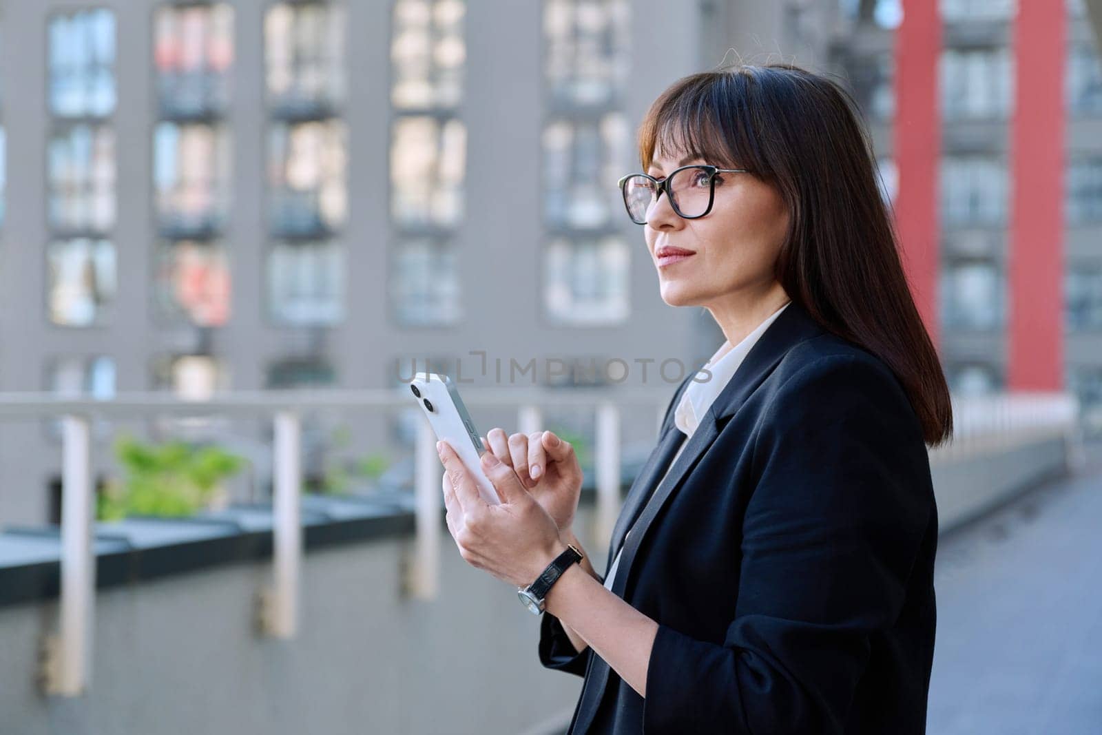 Mature business confident female using smartphone, modern city background. Middle aged successful woman texting using mobile applications apps for business work. Technology internet job, urban style