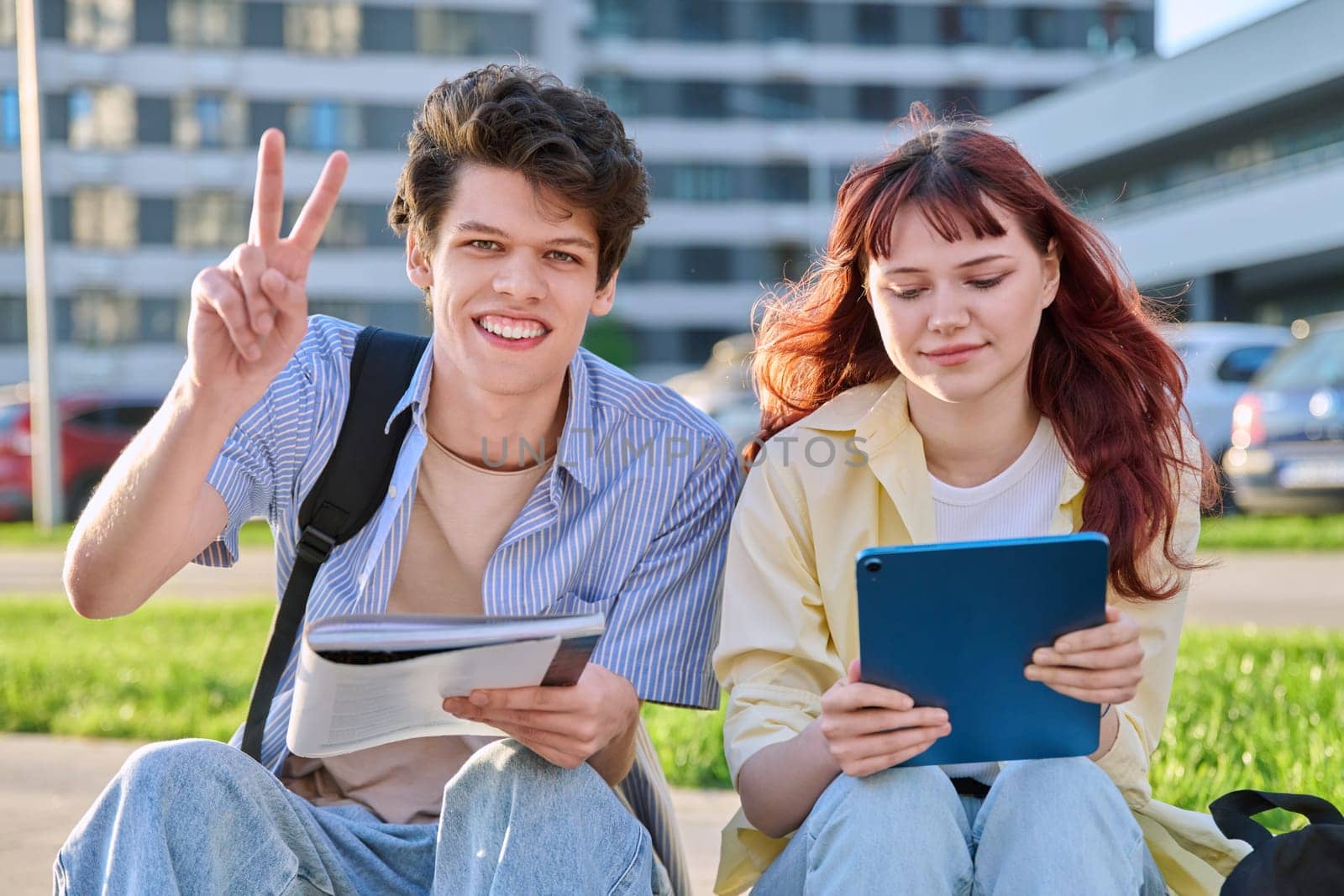 Teenage college students guy and girl talking, using digital tablet, sitting outdoor, urban modern city. Youth 19-20 years old, education, technologies, lifestyle, friendship concept