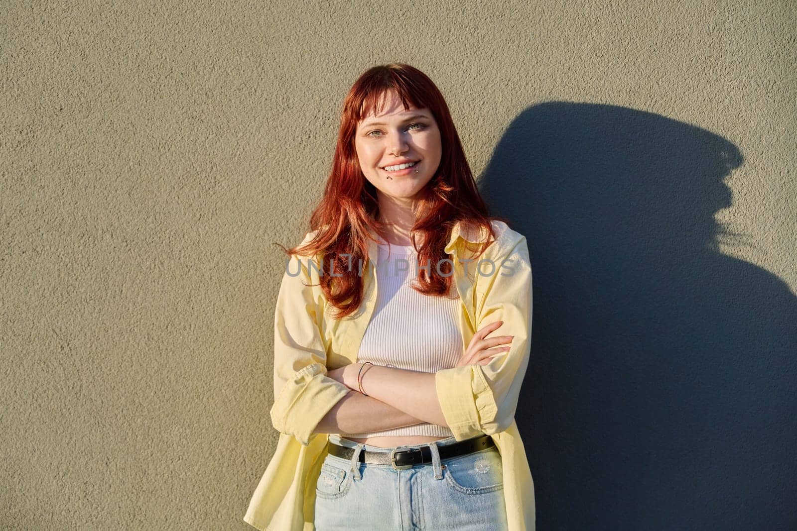 Outdoor portrait of happy confident young hipster female with red hair facial piercing looking at camera with crossed arms sunny gray wall background. Beauty, youth, fashion, joy, happiness, lifestyle