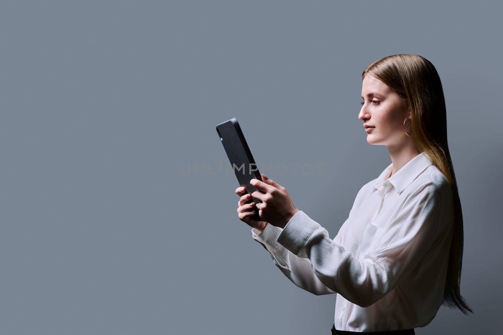 Teenage female student using digital tablet on gray background, profile view. Teenager girl 16,17, 18 years old reading looking at screen. Education Internet technology e-learning educational services