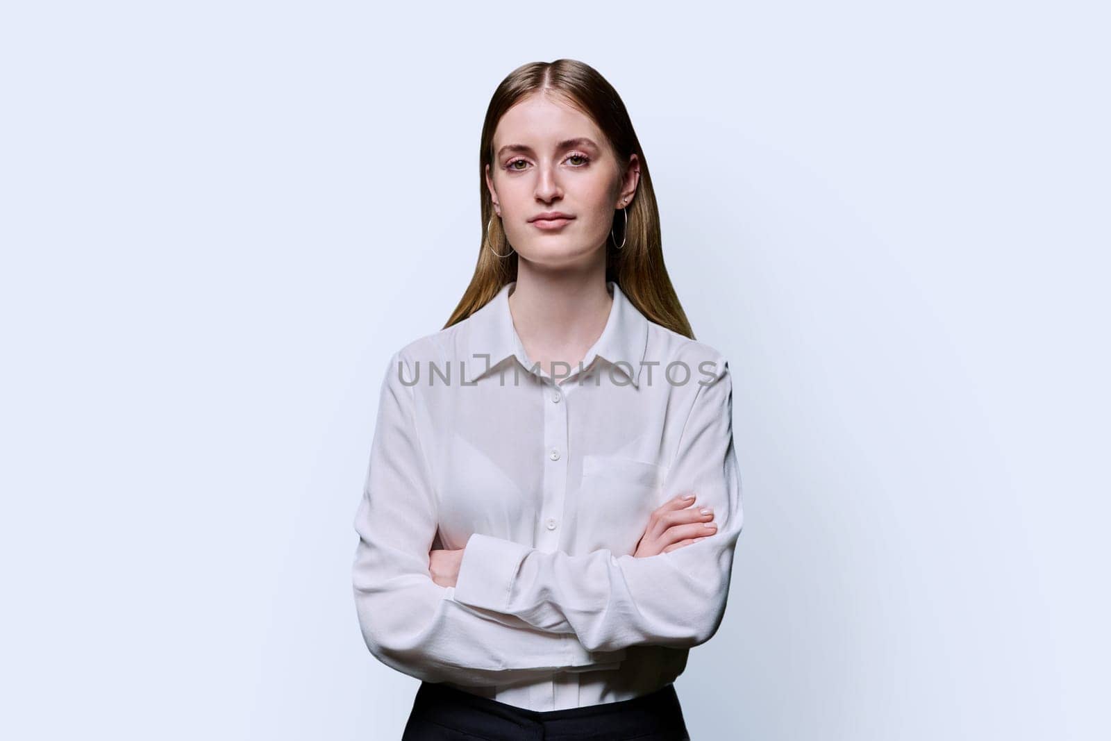 Portrait of young teenage serious female in white shirt with crossed arms on white studio background. Confident beautiful girl college student looking at camera