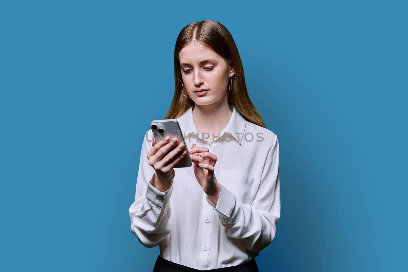 Teenage student girl 16, 17, 18 years old in white shirt holding smartphone in hands, texting looking at screen on blue studio background. Education technology high school college lifestyle youth