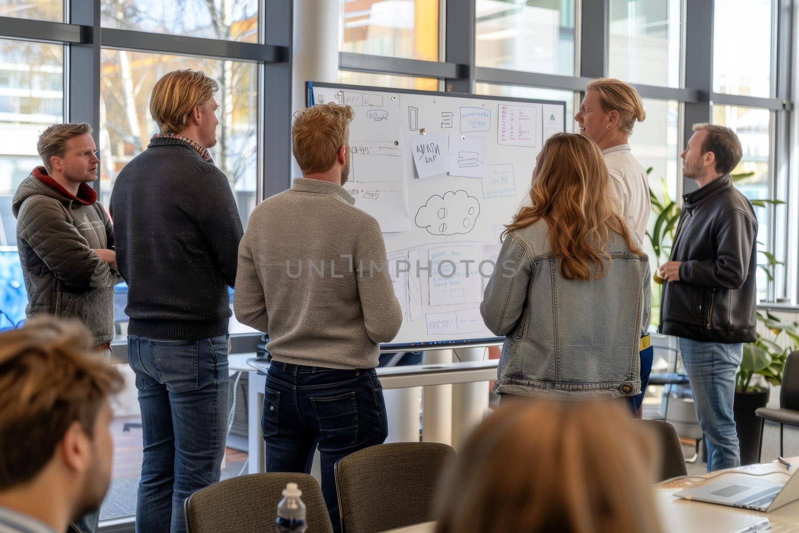 A group of people are standing around a white board with drawings on it. They are discussing ideas and collaborating on a project. Scene is focused and productive