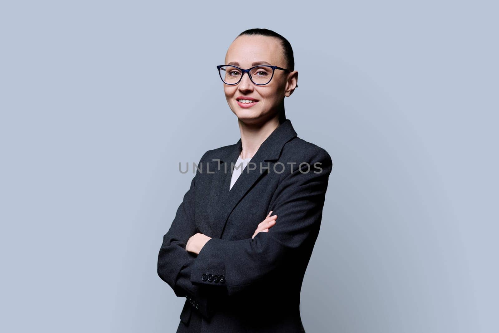 Portrait of happy smiling with teeth business woman in 30s with crossed arms, on grey studio background. Confident female in glasses, suit looking at camera. Business work job career people concept