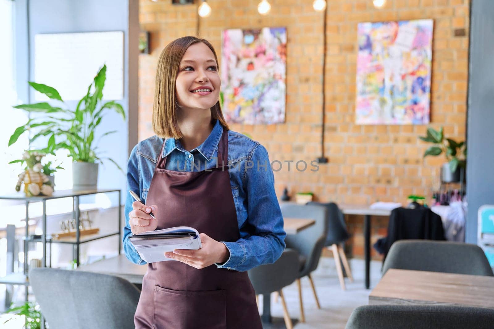 Confident successful young woman service worker owner in apron with working notepad pen in restaurant cafeteria coffee pastry shop interior. Small business staff occupation entrepreneur work