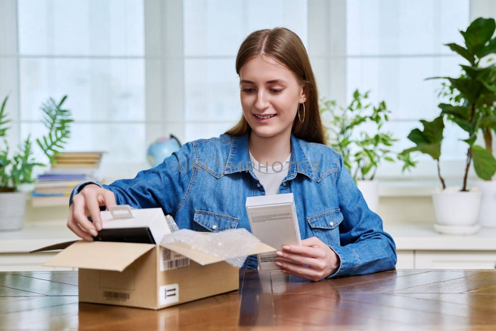 Satisfied young teen female shopper consumer sits at home and unpacks cardboard box with online purchases. Teenager girl unpacking box with new smartphone, headphones. Delivery by mail, online store