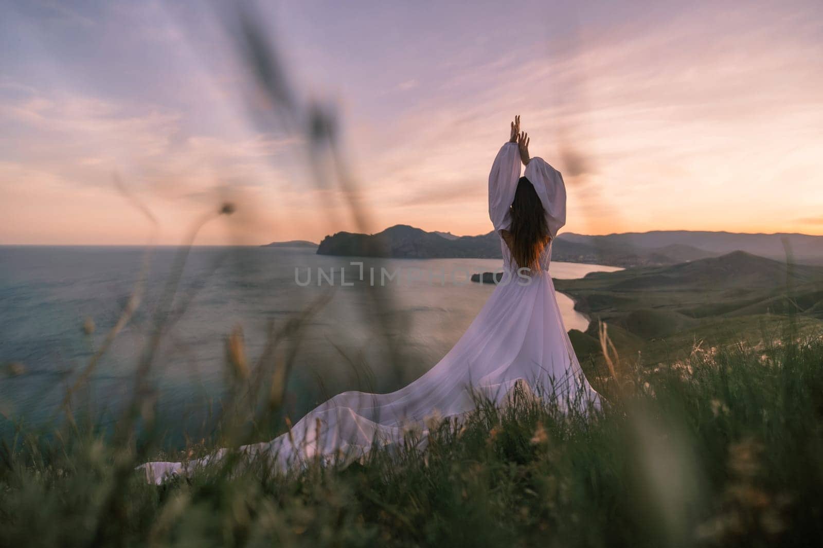A woman in a white dress stands on a hill overlooking the ocean. She is holding her hands up in a yoga pose, and the sky is a beautiful mix of colors. Concept of peace and serenity. by Matiunina