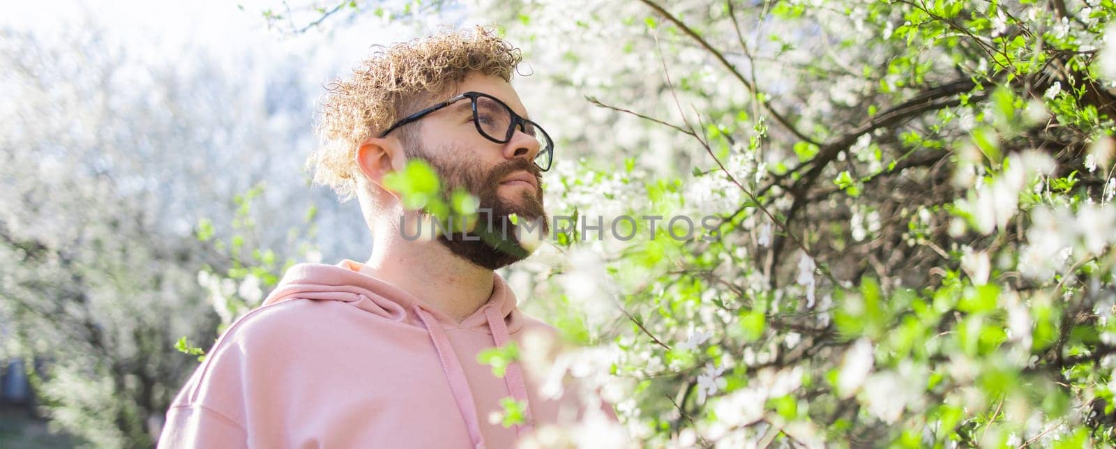 Male bearded guy standing under branches with flowers of blooming almond or cherry tree in spring garden. Spring blossom. Copy space
