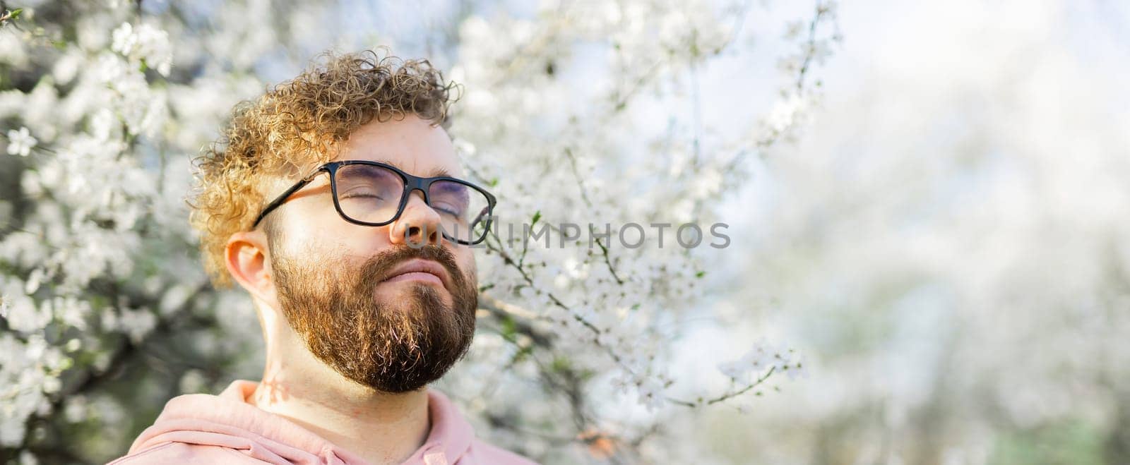 Banner Male bearded man standing under branches with flowers of blooming almond or cherry tree in spring garden. Spring blossom. Copy space by Satura86