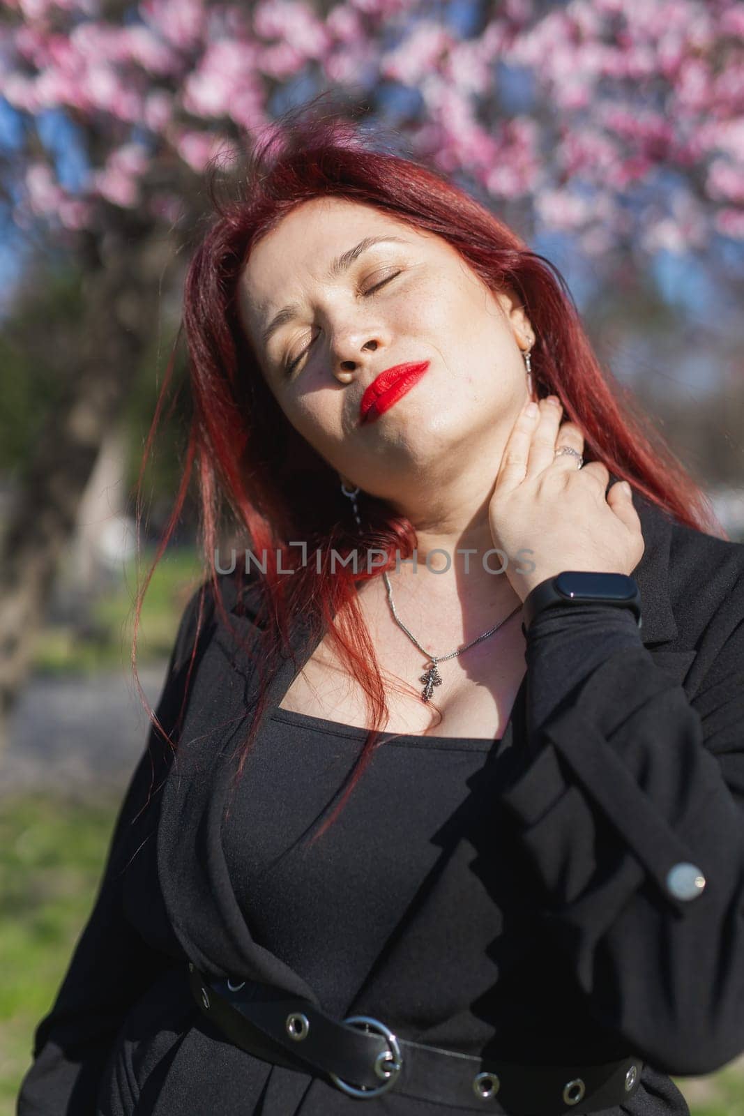 Fashion outdoor photo of beautiful woman with red curly hair in elegant suit posing in spring flowering park with blooming cherry tree. Copy space and empty place for advertising text.