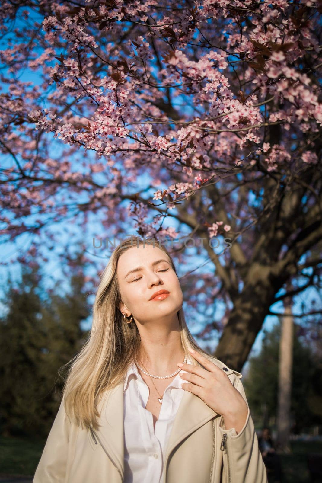 Fashion outdoor photo of beautiful woman with blond hair in elegant suit posing in spring flowering park with blooming cherry tree. Copy space and empty place for advertising text by Satura86