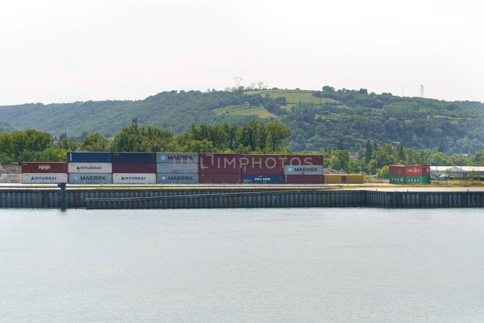 Lyon, France - May 30, 2023: Stacked shipping containers in varying colors beside a calm river, with a green hill in the background under a clear sky.