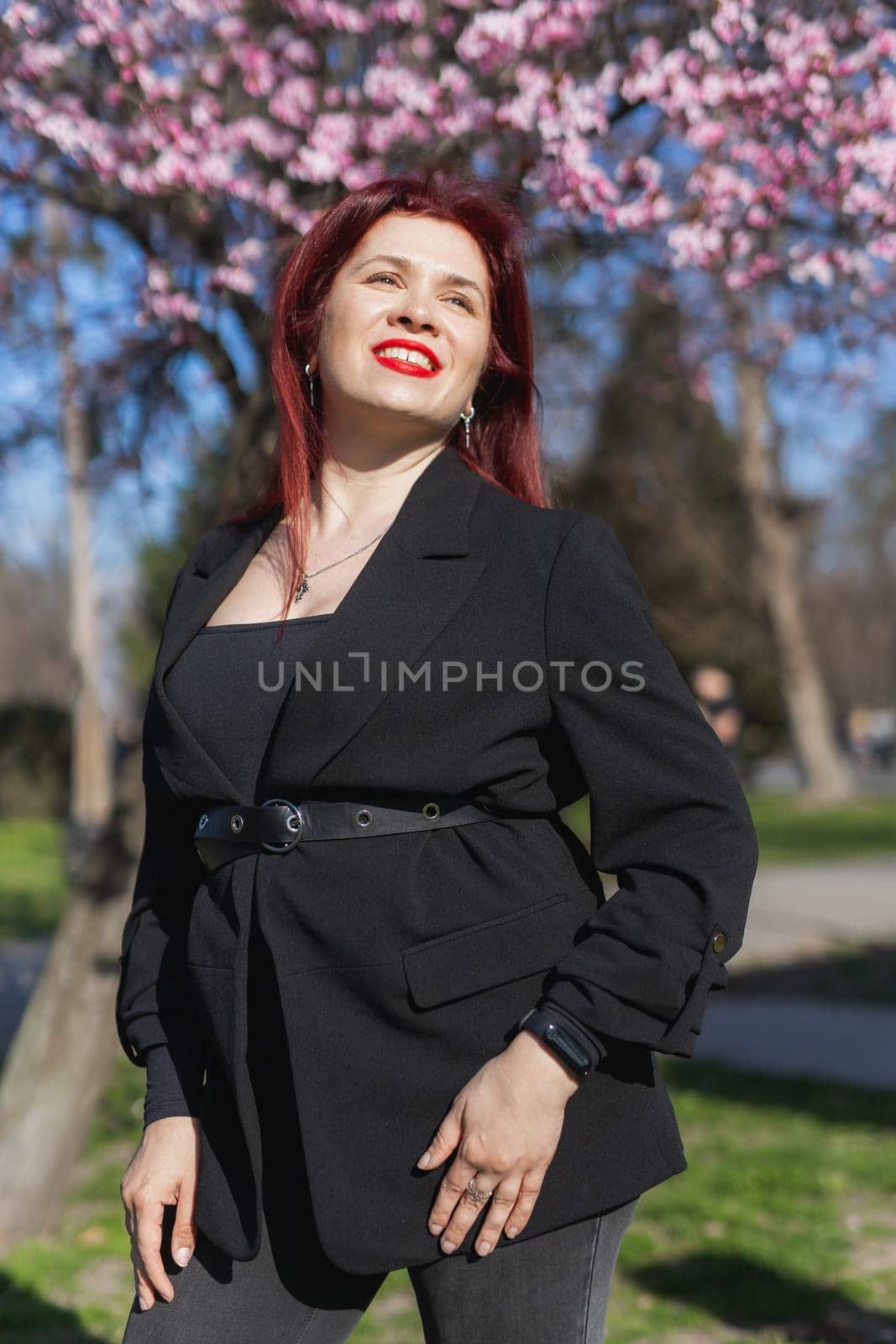 Fashion outdoor photo of beautiful woman with red curly hair in elegant suit posing in spring flowering park with blooming cherry tree. Copy space and empty place for advertising text by Satura86