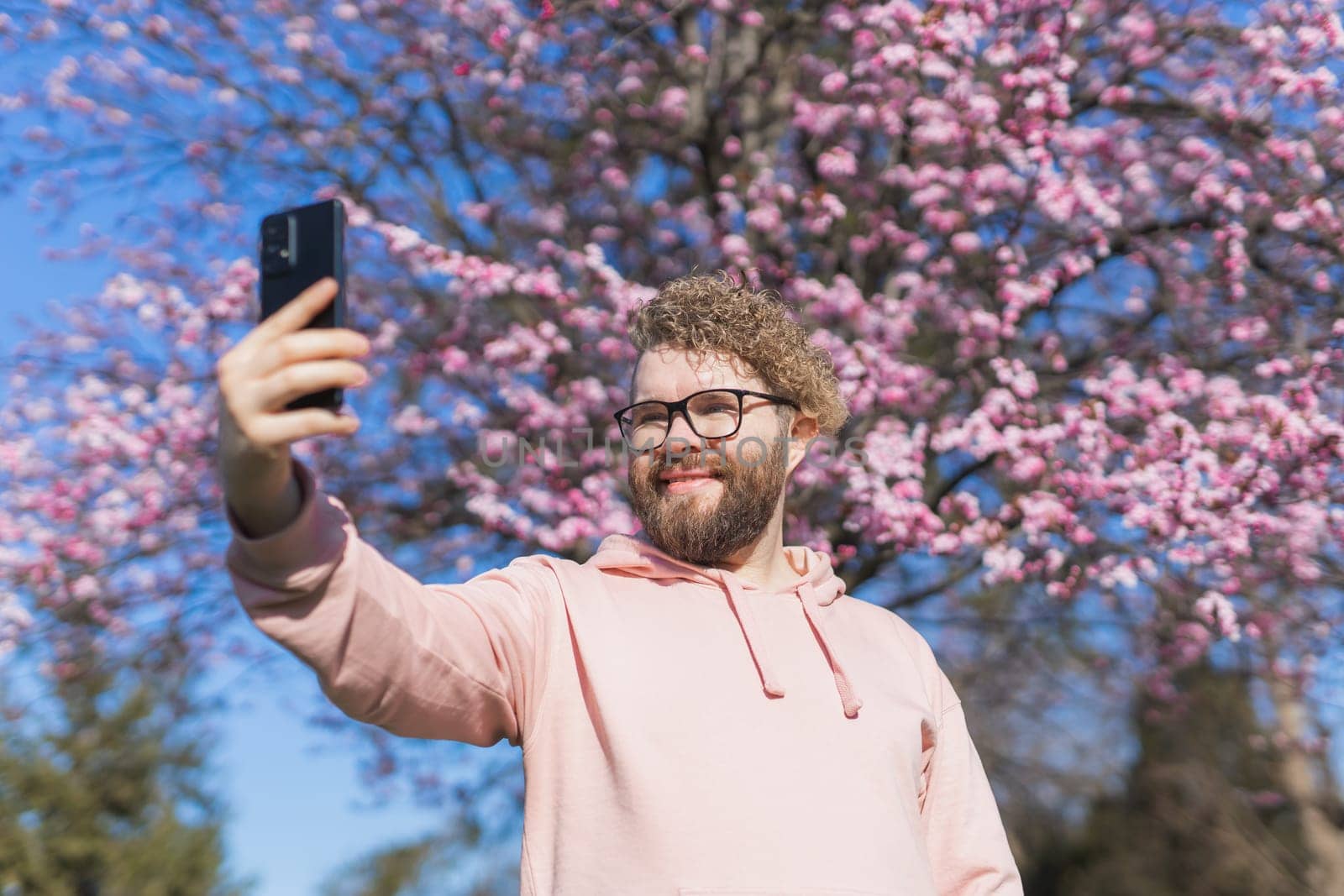 Spring day. Bearded man in pink shirt talking by phone. Spring pink sakura blossom. Handsome young man with smartphone. Fashionable man in trendy glasses. Bearded stylish man. Male fashion.