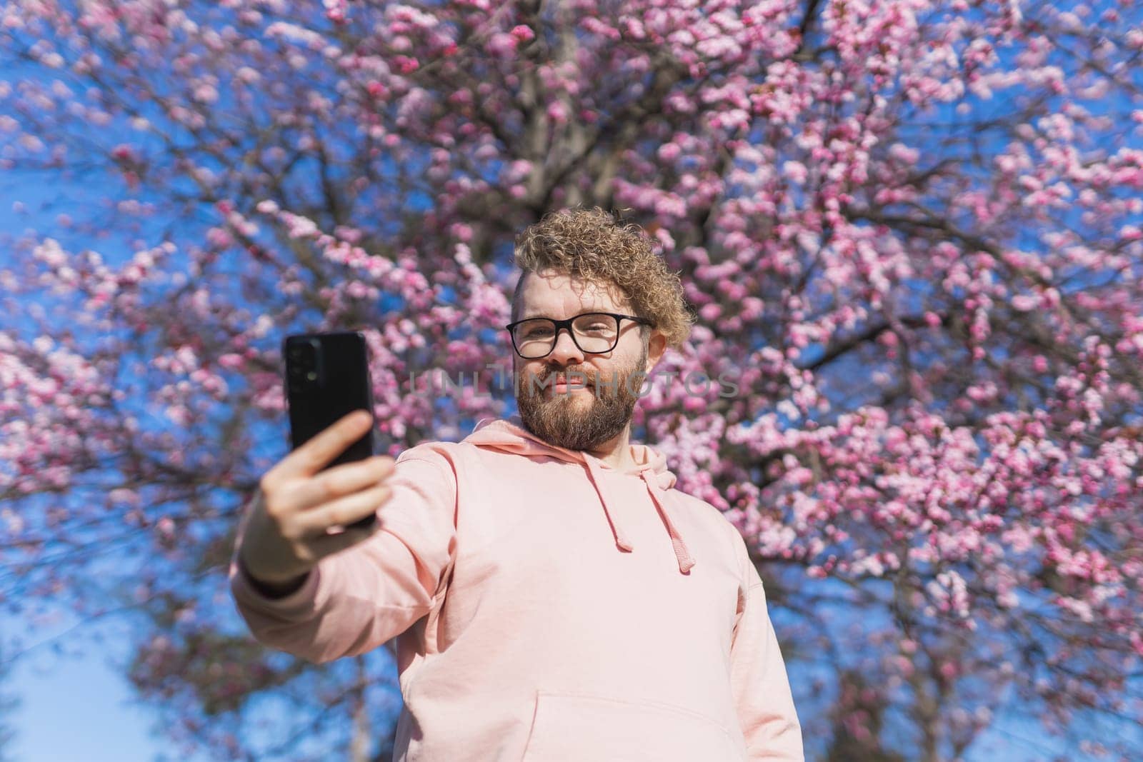 Happy curly man takes selfie against backdrop of flowering tree in spring for his internet communications. Weekend and social networks