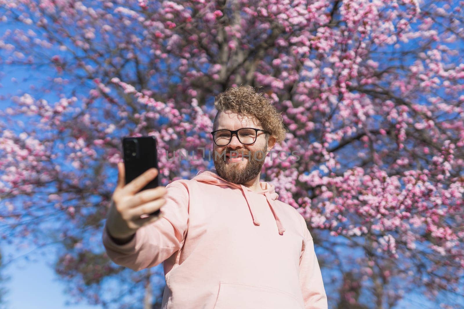 Spring day. Bearded man in pink shirt talking by phone. Spring pink sakura blossom. Handsome young man with smartphone. Fashionable man in trendy glasses. Bearded stylish man. Male fashion.