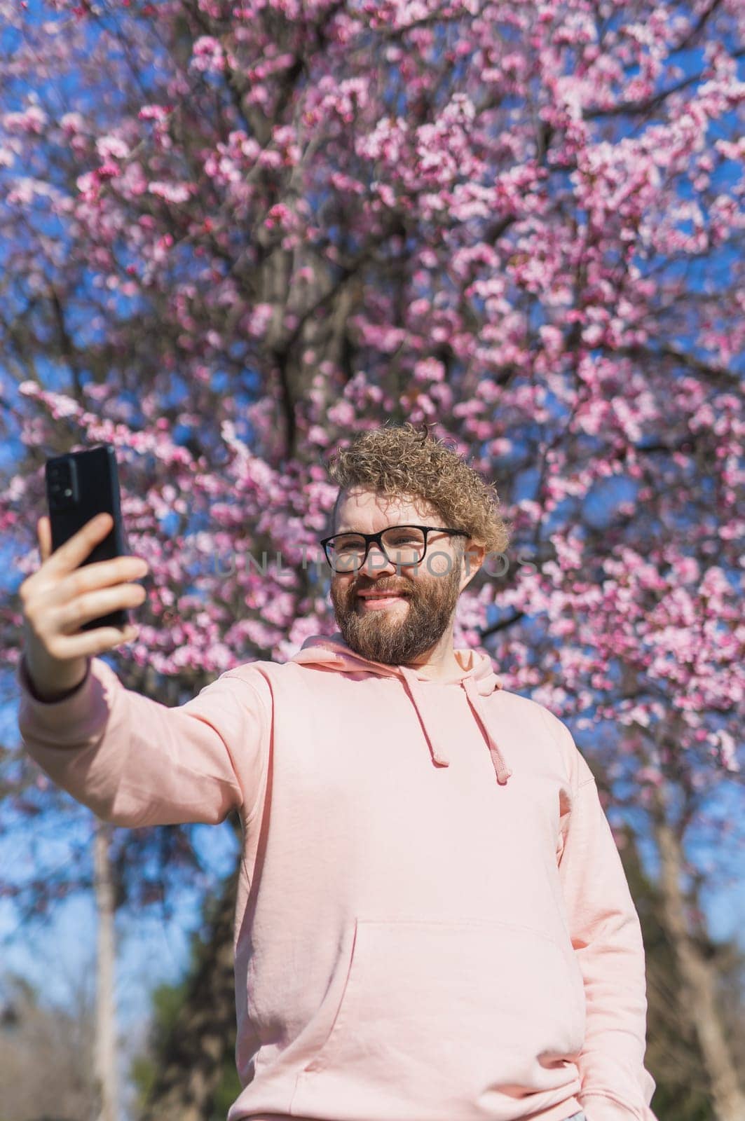 Happy curly man takes selfie against backdrop of flowering tree in spring for his internet communications. Weekend and social networks