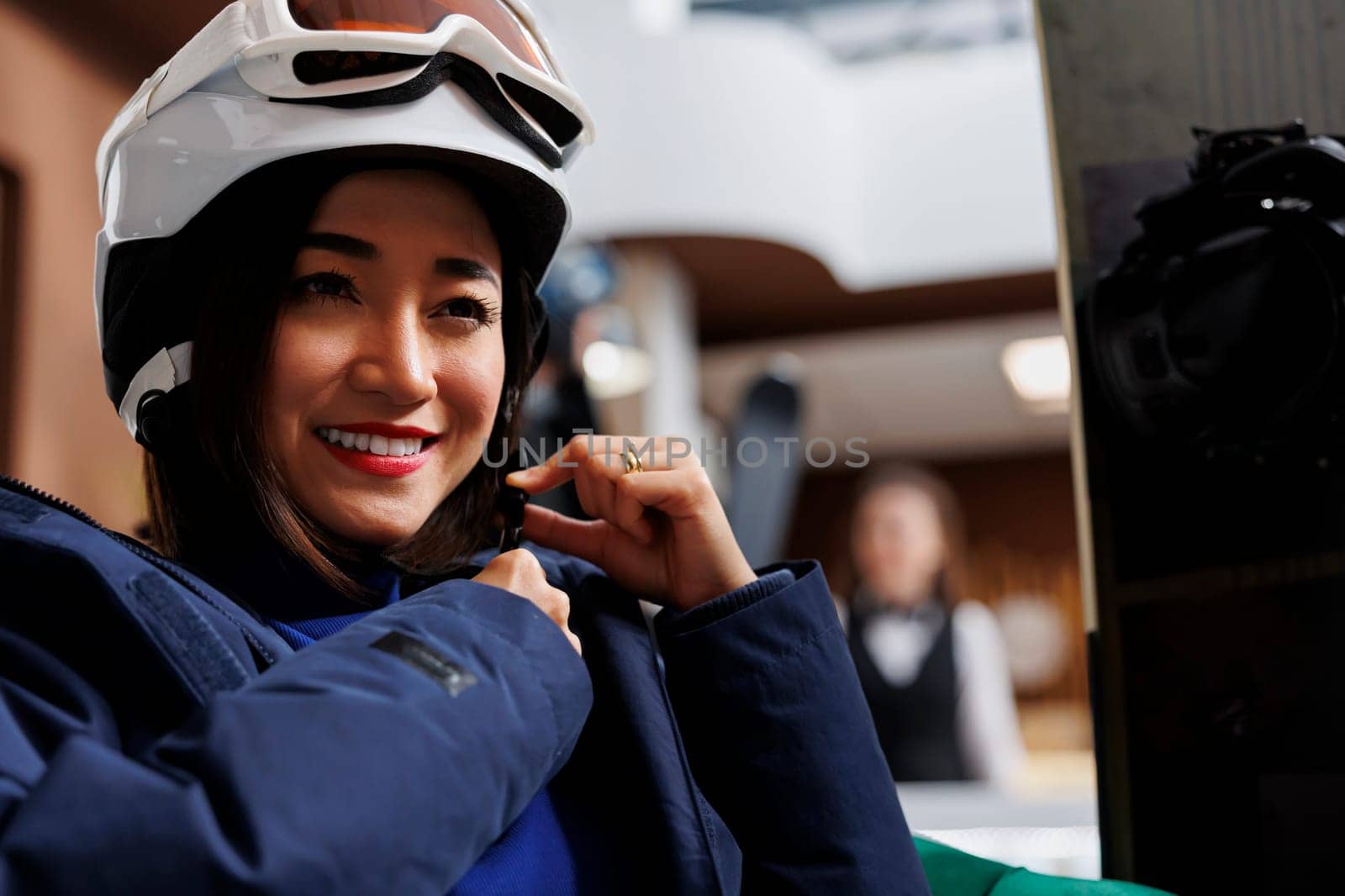 Asian woman securing band of winter helmet while sitting in hotel foyer and sporting a winter jacket. Woman makes safety modification to snow headgear for snowboarding on slopes of ski resort.