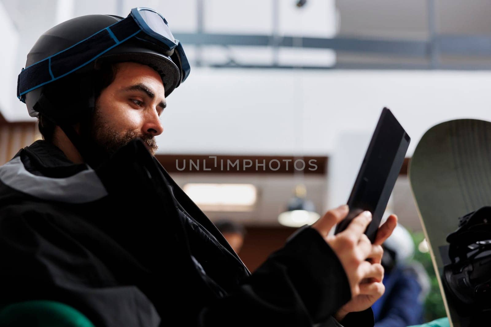 Close-up view of young man dressed in winter jacket browsing the internet on a digital device. Image showing male traveler with snowboarding gear engrossed in his tablet searching for snowy slopes.