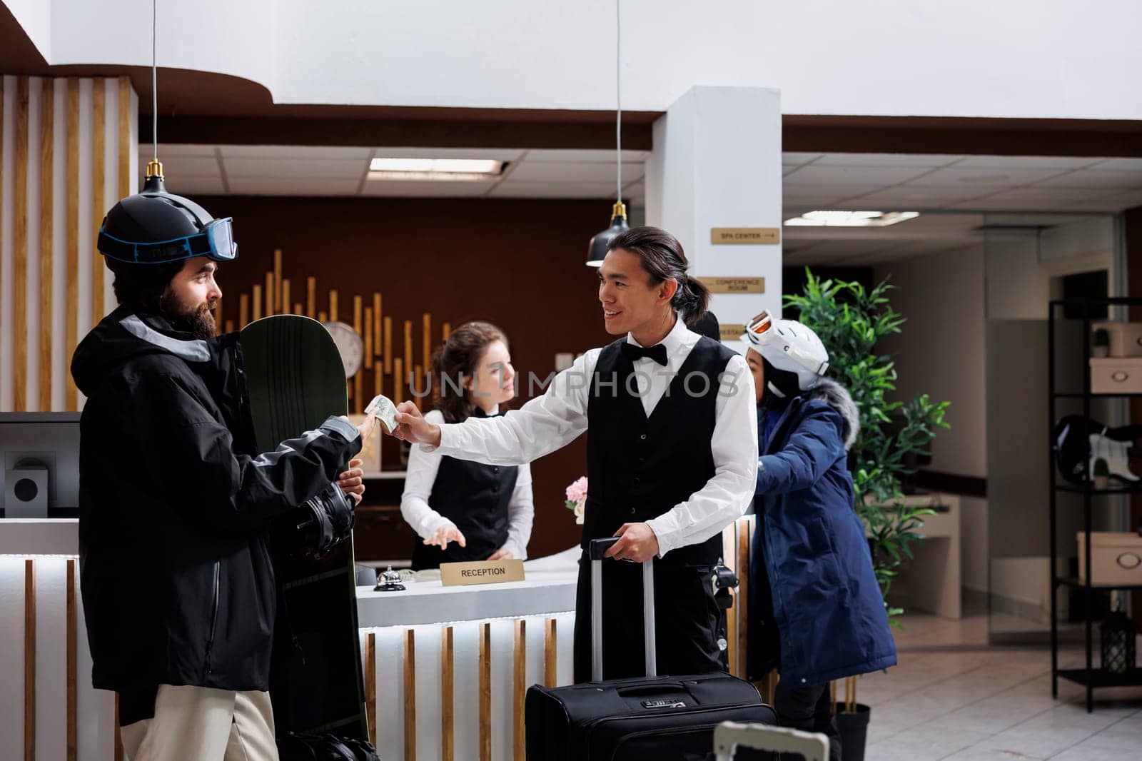 Tourists check in at ski mountain resort with luggage and snowboard equipment. Receptionist assists with reservations, male guest wearing winter clothing gives cash tip to staff in hotel lobby.