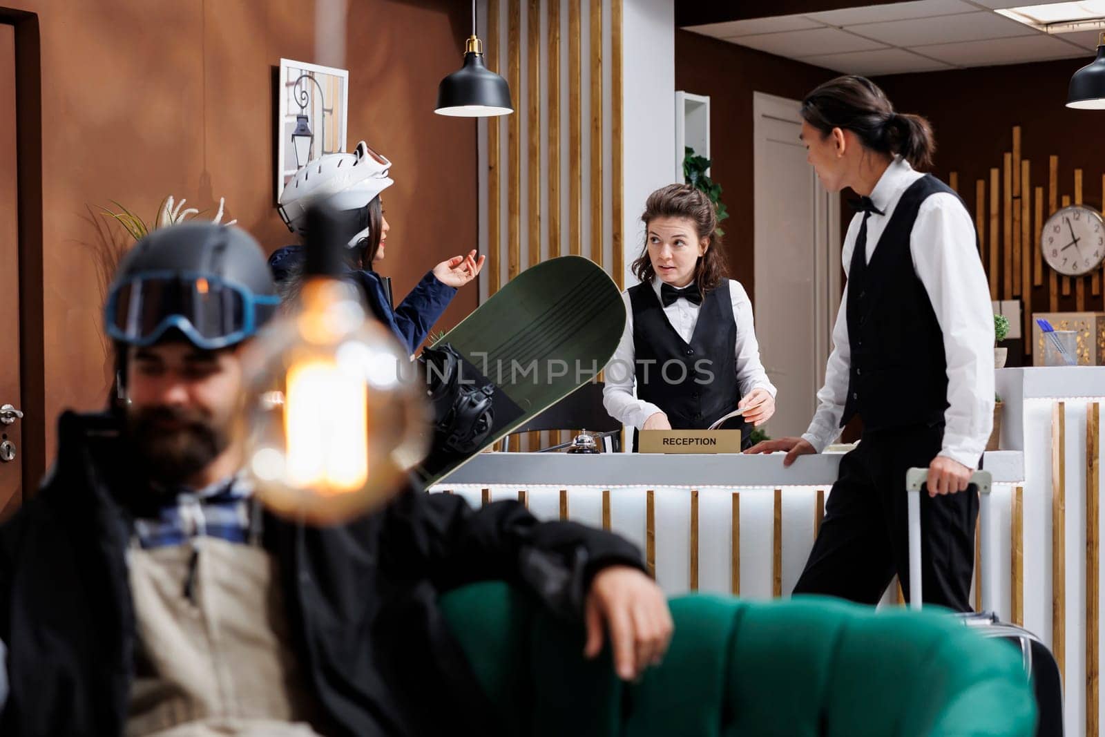 Arriving at hotel reception staff provides excellent service female guest while man sits on sofa waiting for hotel room. Winter gear and ski equipment hints a thrilling winter holiday at ski resort.