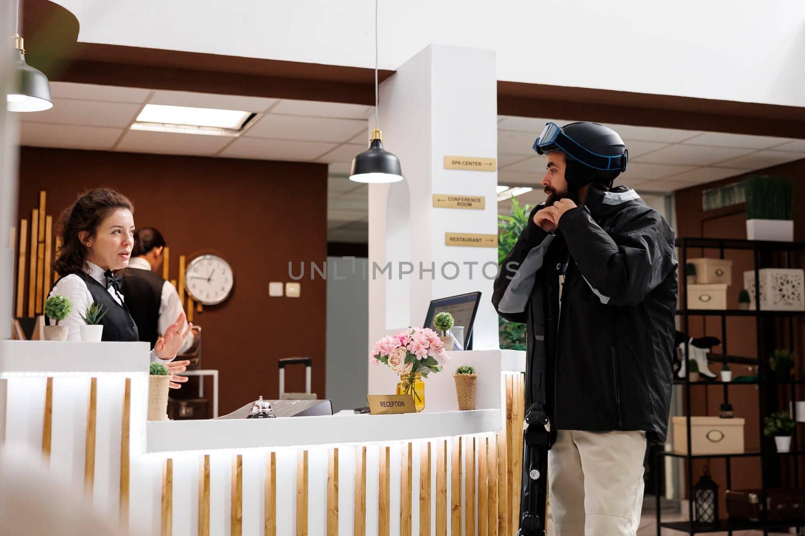 Caucasian man checking in at hotel reception for winter vacation at ski resort. Friendly female staff welcomes customer at front desk in cozy lounge area. Suitcases and ski gear in tow.