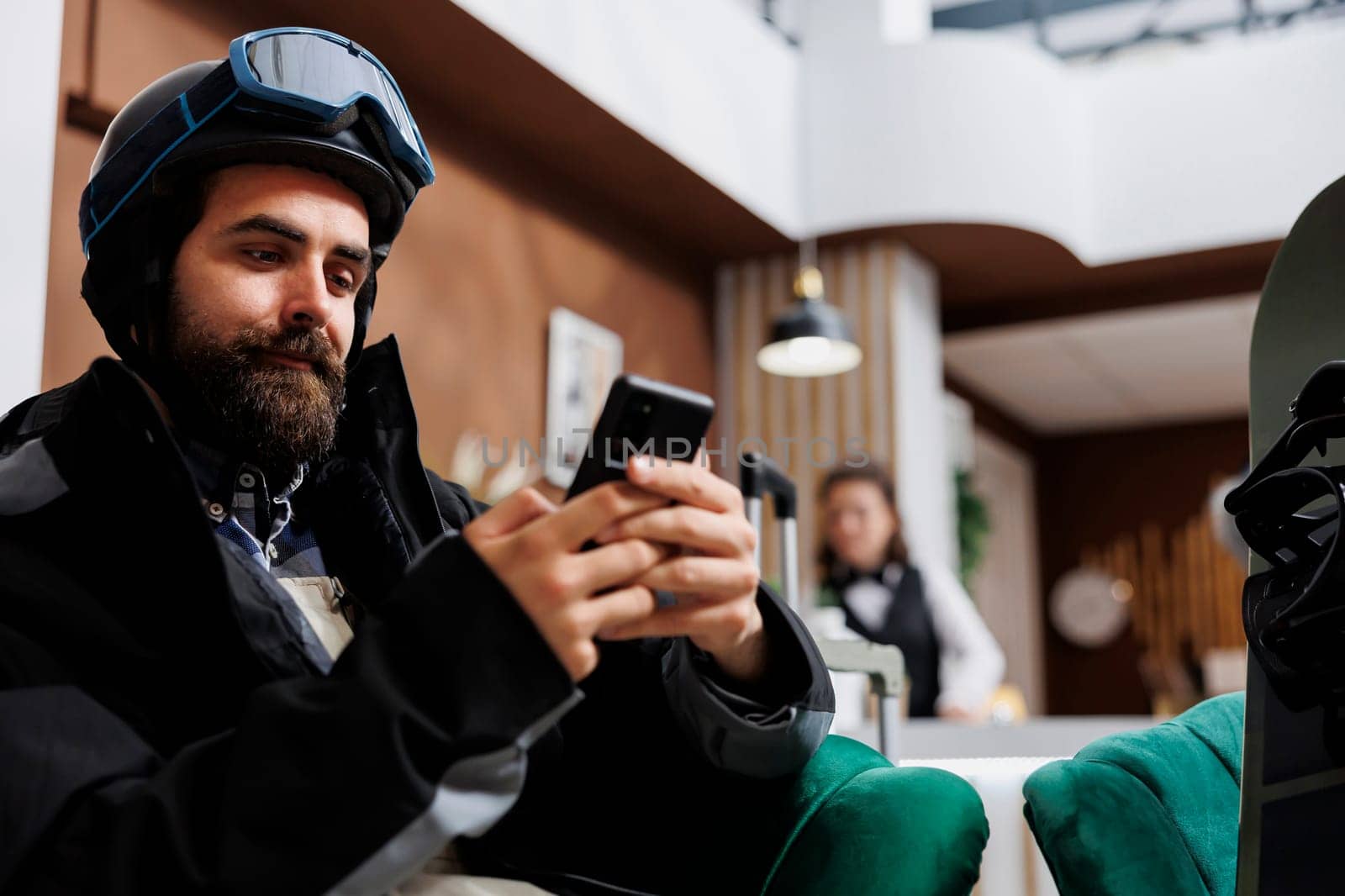 During winter holiday young man uses mobile phone in cozy lounge area. Male guest sitting on sofa with digital smart device and snowboard wearing winter jacket, ski goggles and helmet.
