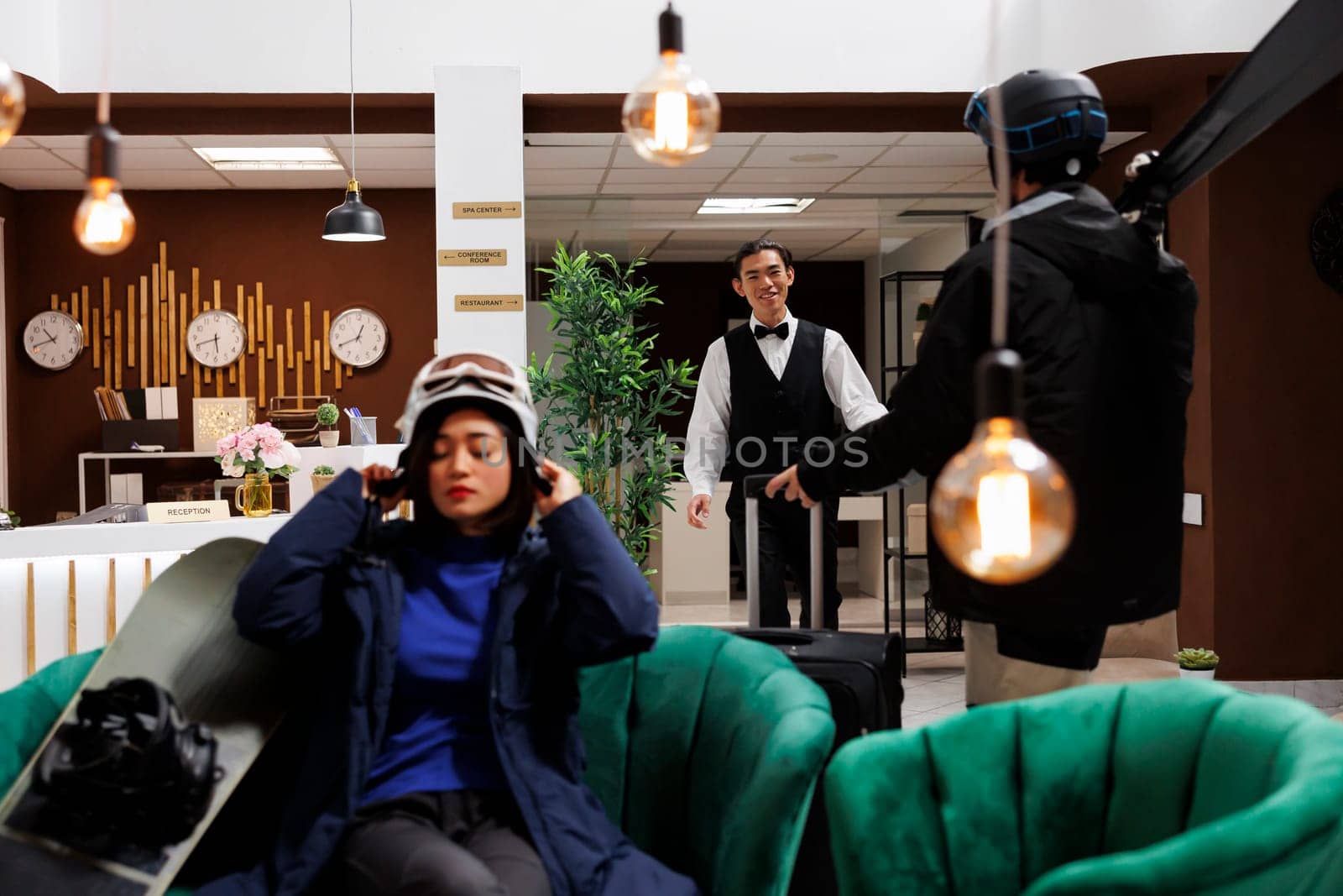 Male traveler arrives at cozy hotel lobby for skiing winter getaway being welcomed by asian employee. Young woman adjusting helmet for snowboarding activity at luxury mountain resort.