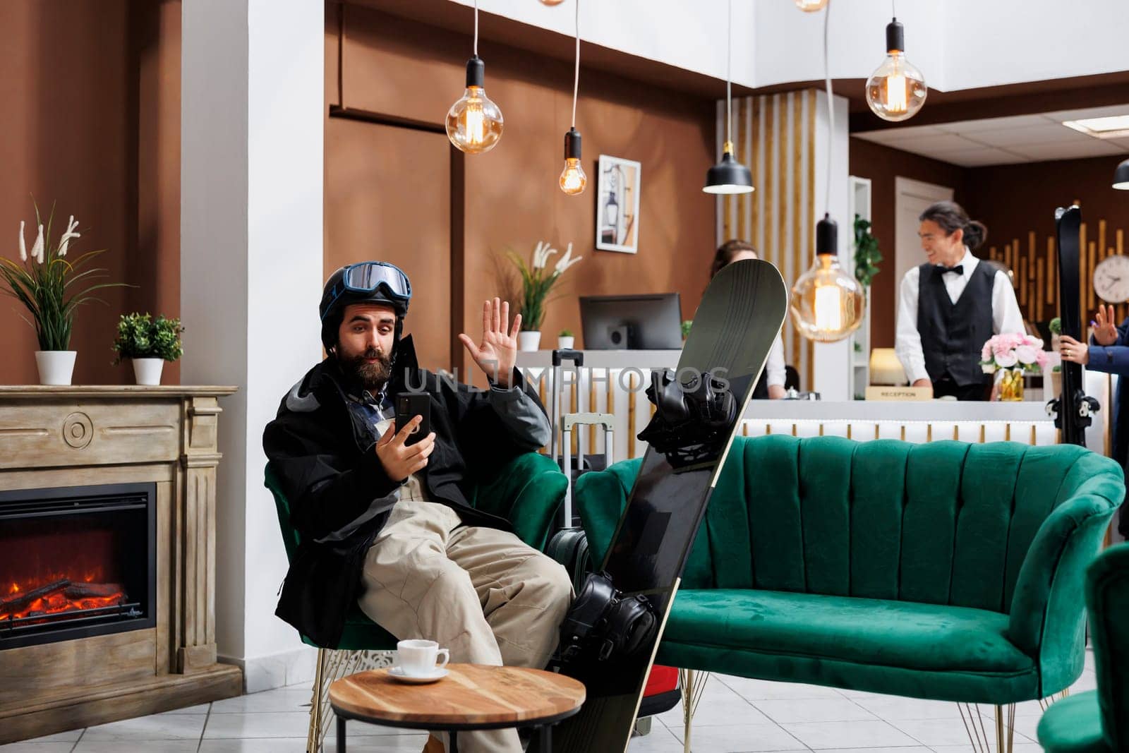 A male tourist in hotel lobby, using his smartphone for videoconferencing while waiting. Snowboarding gear and ski goggles suggest stay at ski resort. Cozy and tech-savvy ambiance for travelers.