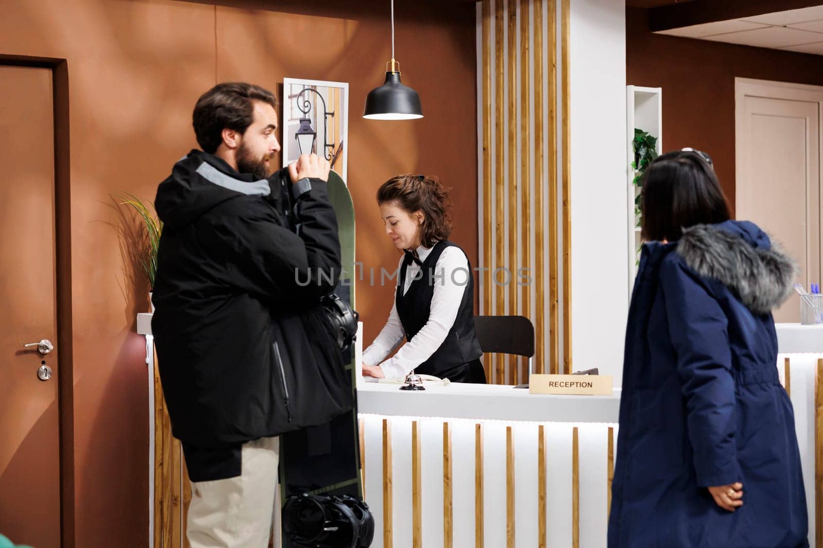 Youthful man and woman at front desk being assisted with check-in by friendly hotel receptionist. Male tourist holding snowboard while female speaks with employee in resort lounge area.