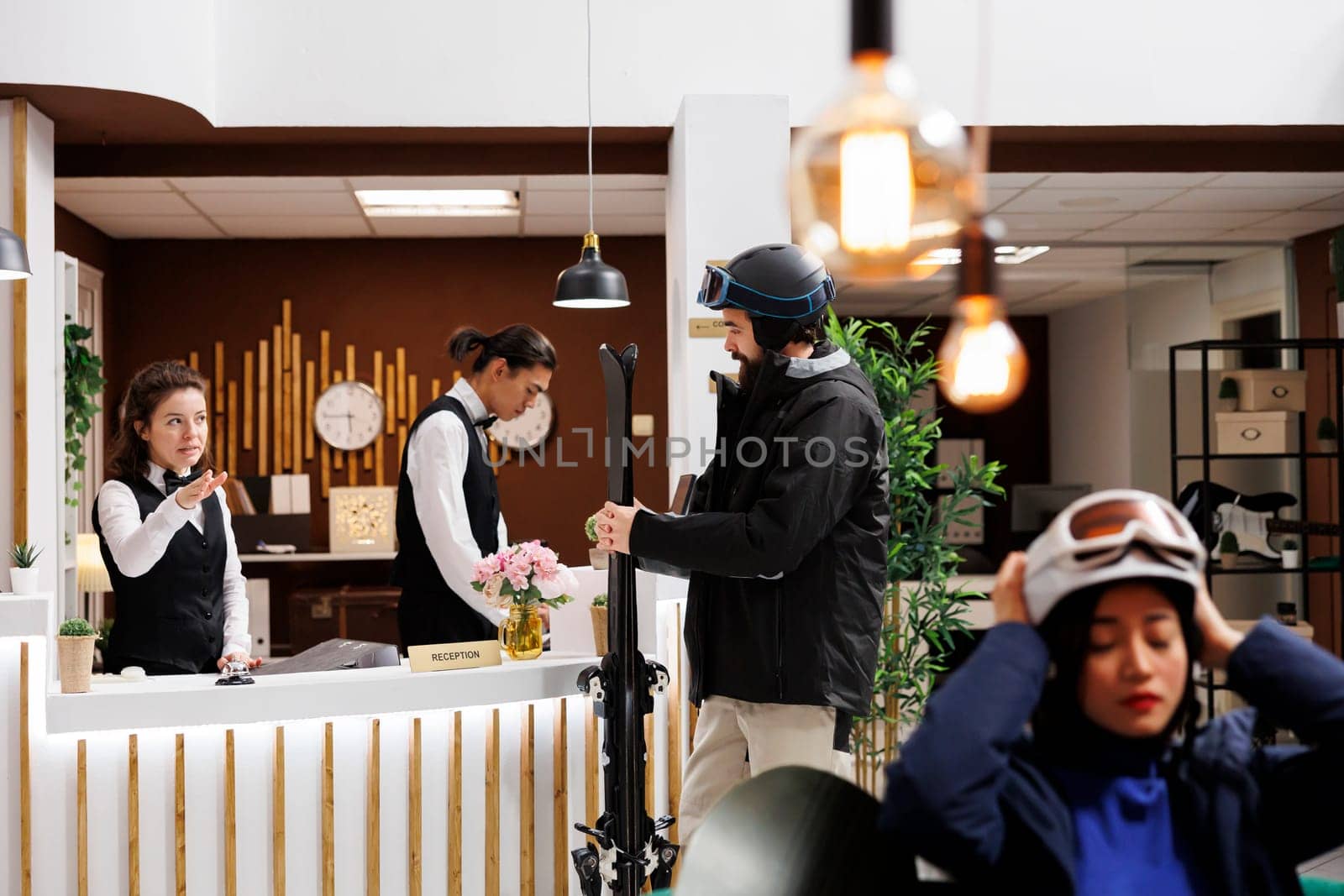 Female guest with helmet waits in luxurious lounge area ready for winter vacation at resort. Excitement fills the air as male traveler holding ski equipment checks in at hotel reception front desk.