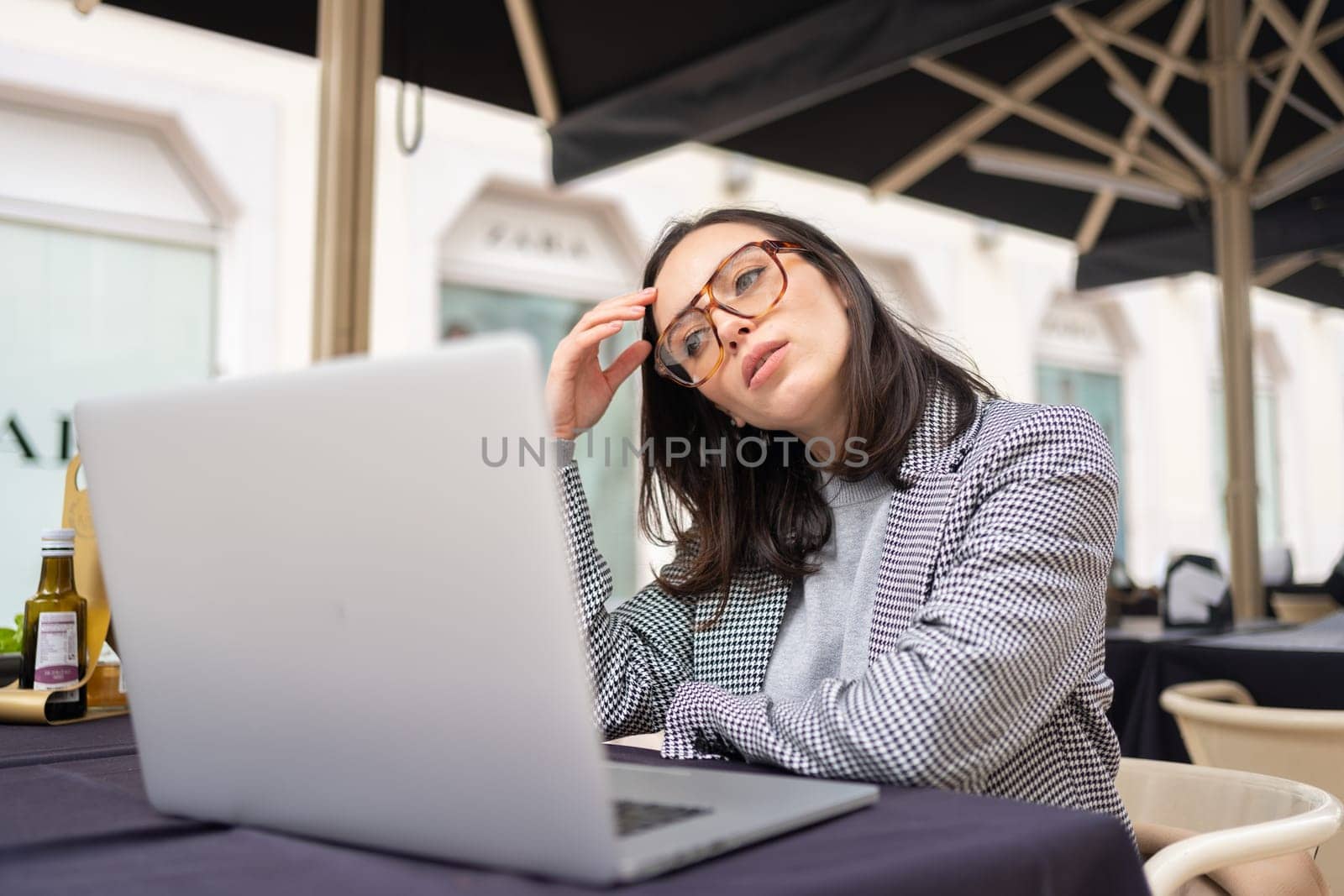 Tired business woman in stress works at laptop while sitting at cafe table on street and holds her hand on her temples, migraine attack. Freelance overworked