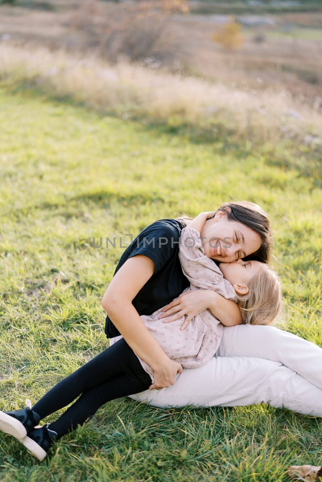 Little girl kisses her mother on the cheek while lying in her arms on a green meadow by Nadtochiy