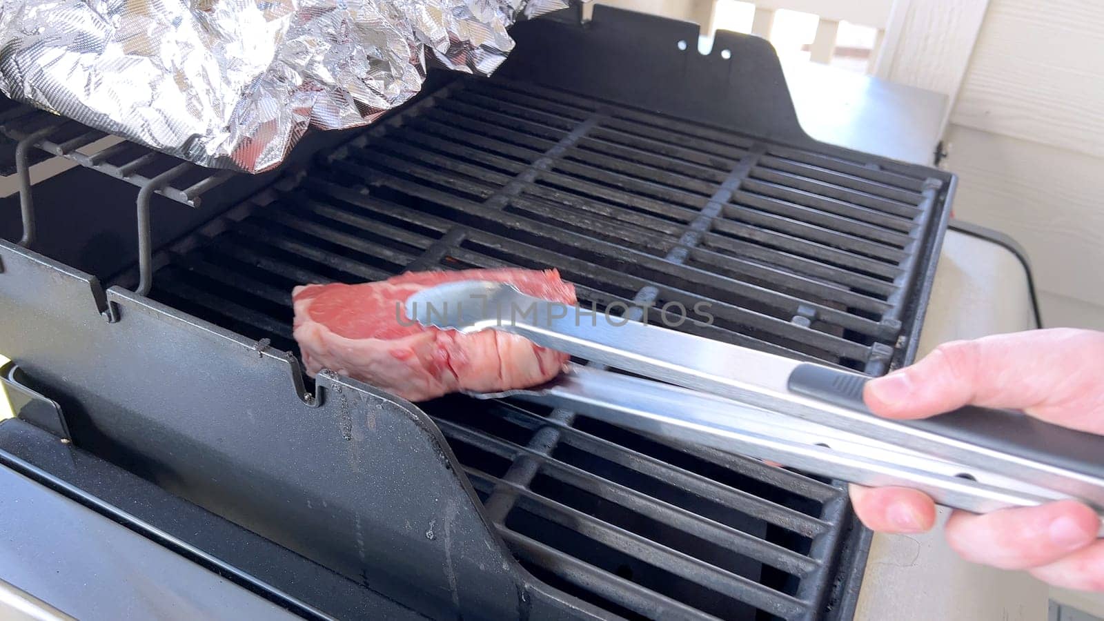 This image showcases the art of grilling, featuring three thick steaks cooking on a barbecue grill, with a row of foil-wrapped corn on the cob above, capturing a typical scene of a hearty outdoor meal preparation.