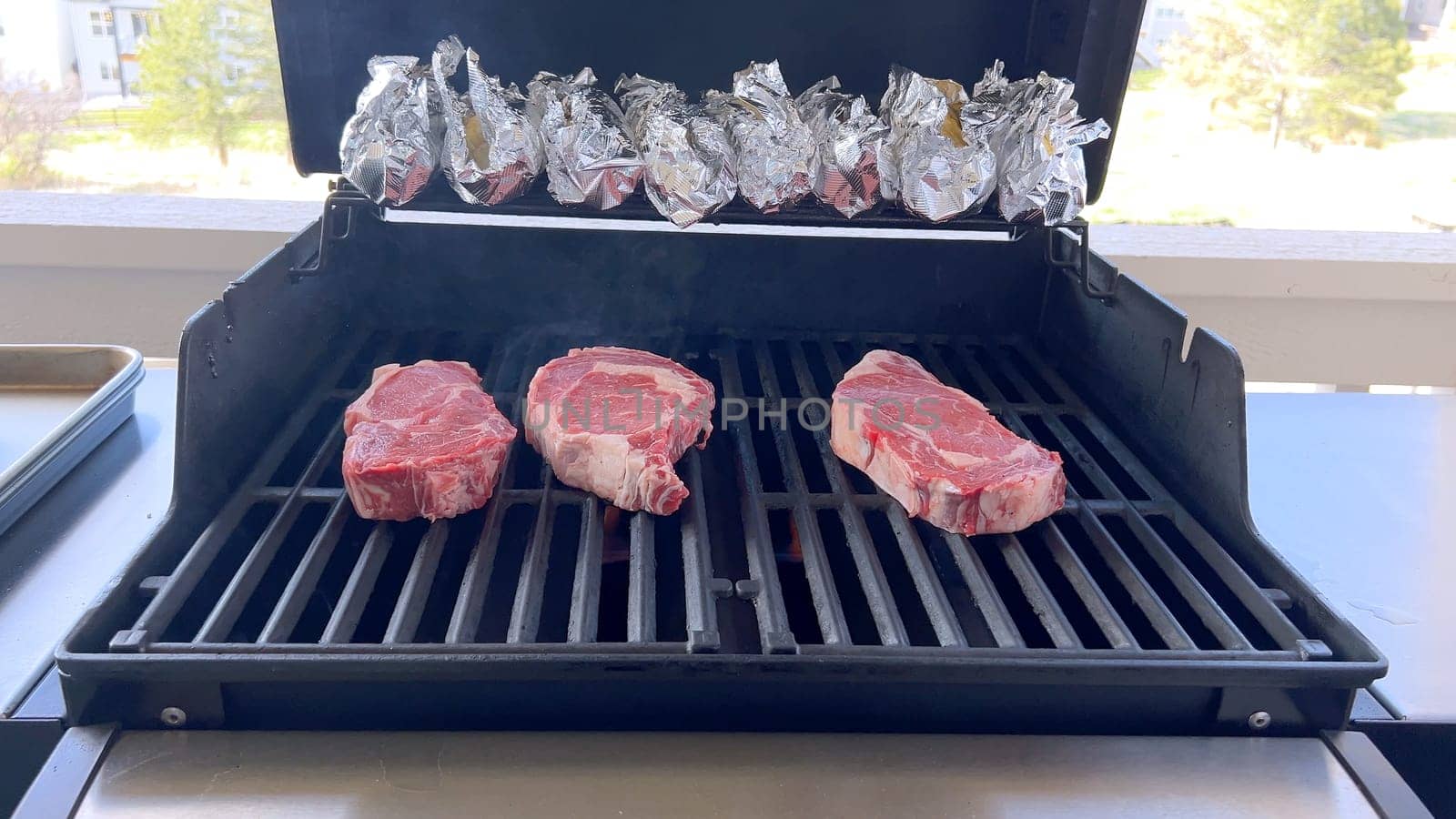 This image showcases the art of grilling, featuring three thick steaks cooking on a barbecue grill, with a row of foil-wrapped corn on the cob above, capturing a typical scene of a hearty outdoor meal preparation.