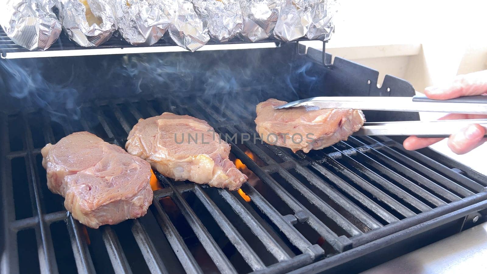 A person uses tongs to flip juicy steaks on a grill, with flames licking the meat, captured in a moment of intense heat and sizzle, showcasing a classic outdoor cooking experience.