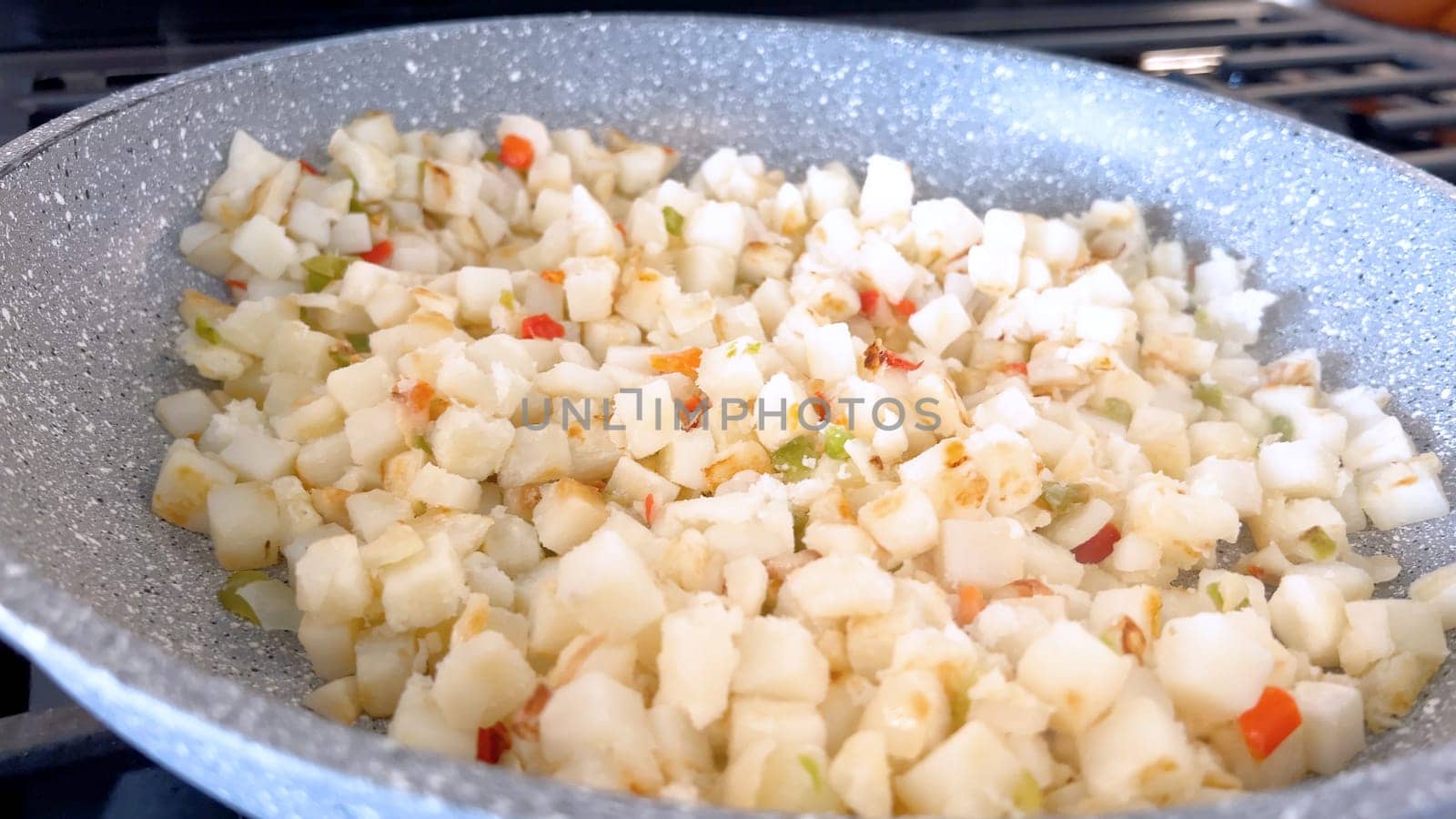 A frying pan on a gas stove sizzles with diced potatoes and colorful bits of red and green peppers, showcasing a delicious and simple home-cooked side dish in the making.