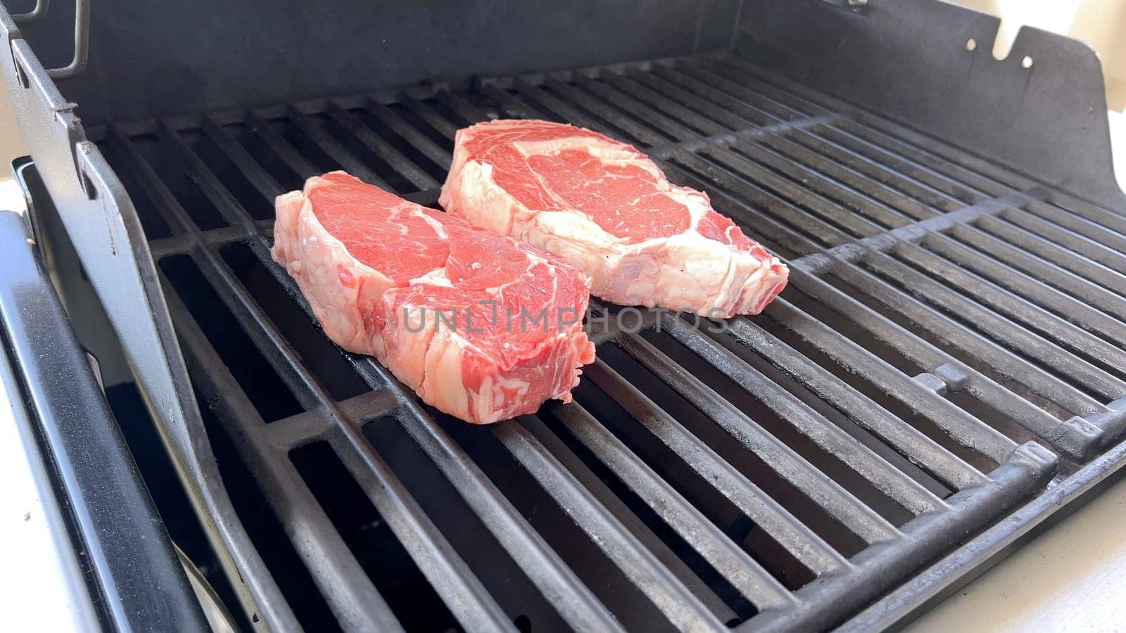This image showcases the art of grilling, featuring three thick steaks cooking on a barbecue grill, with a row of foil-wrapped corn on the cob above, capturing a typical scene of a hearty outdoor meal preparation.