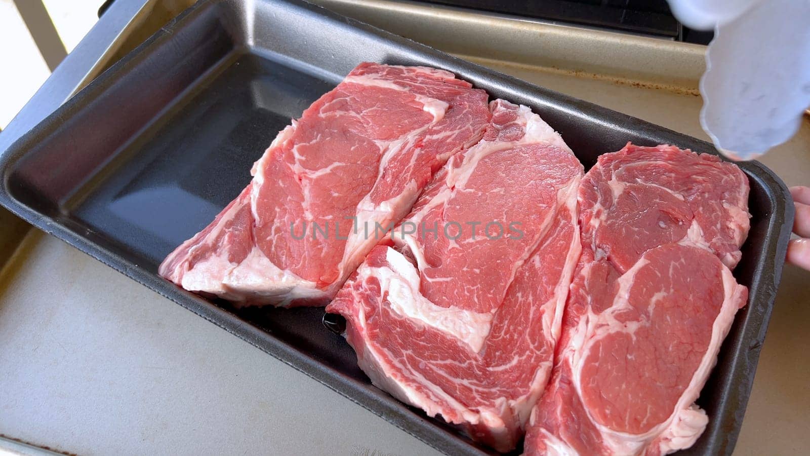 This image showcases the art of grilling, featuring three thick steaks cooking on a barbecue grill, with a row of foil-wrapped corn on the cob above, capturing a typical scene of a hearty outdoor meal preparation.