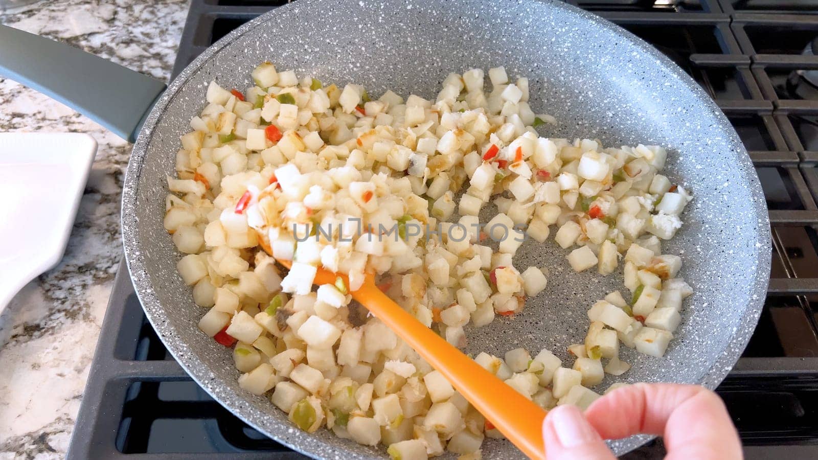 A frying pan on a gas stove sizzles with diced potatoes and colorful bits of red and green peppers, showcasing a delicious and simple home-cooked side dish in the making.