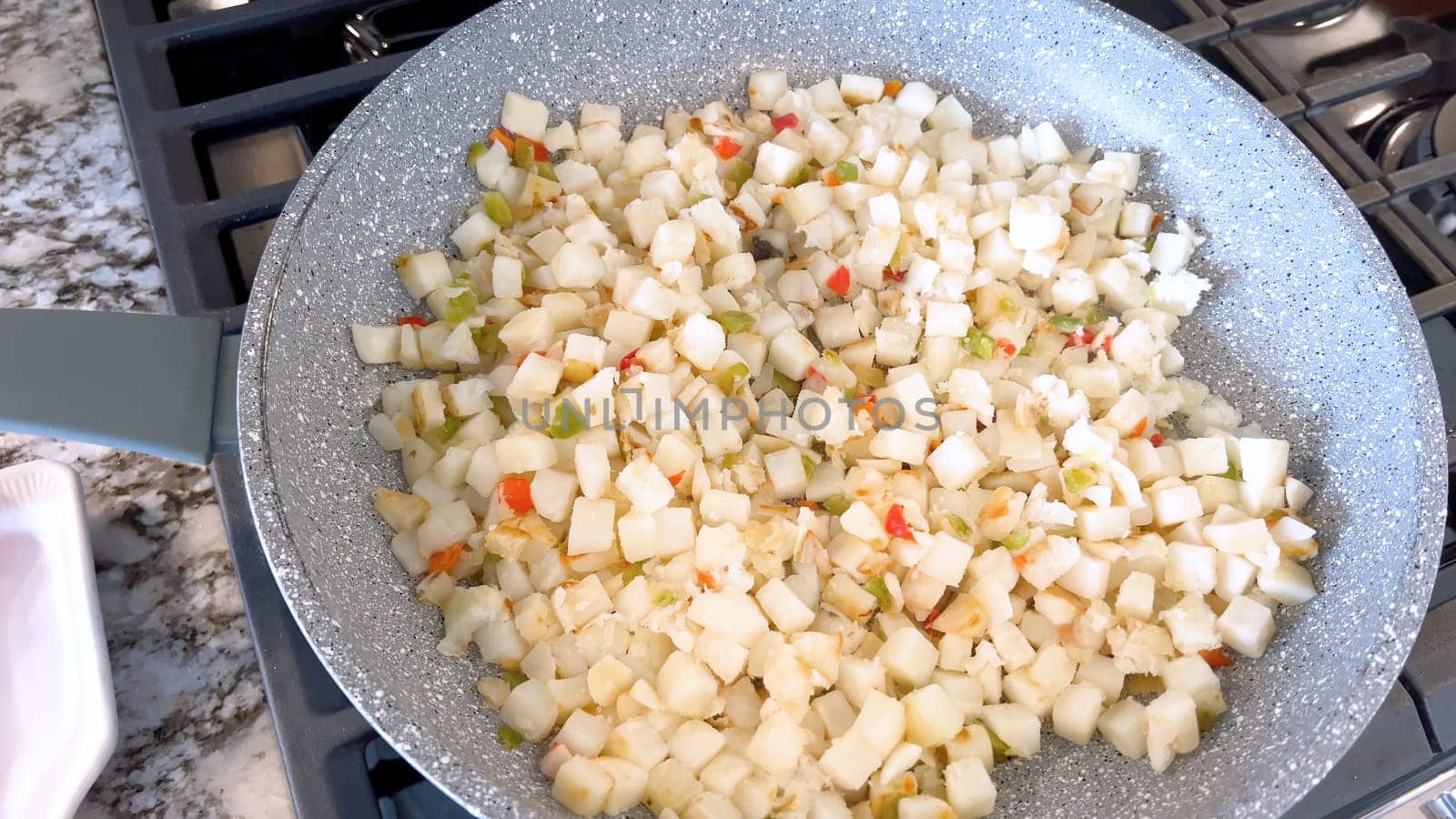 A frying pan on a gas stove sizzles with diced potatoes and colorful bits of red and green peppers, showcasing a delicious and simple home-cooked side dish in the making.