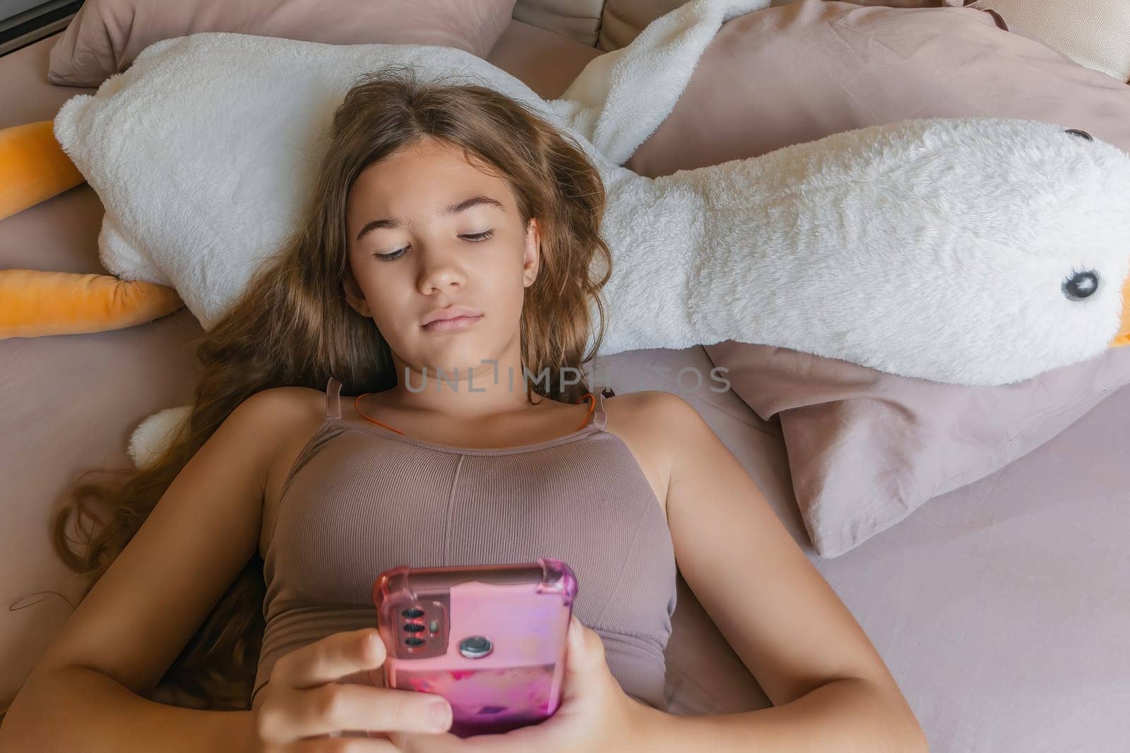 A girl is laying on a bed with a pink phone in her hand. She is looking at the phone and she is focused on something on the screen. by Matiunina