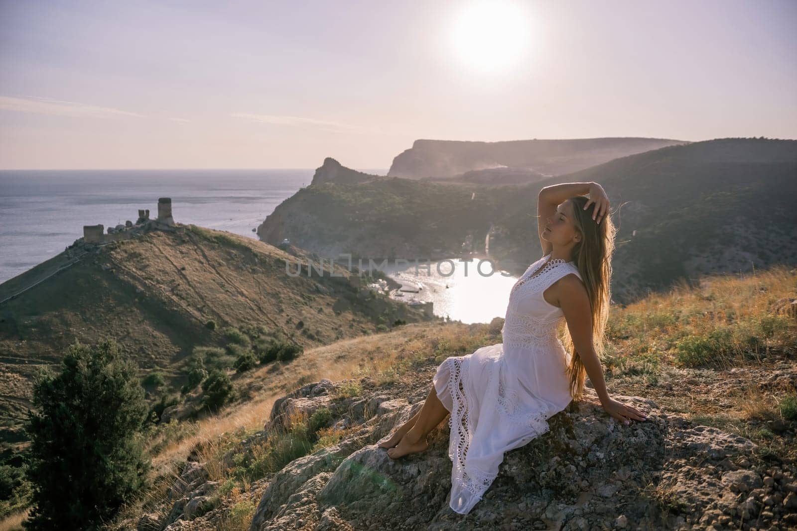 A woman in a white dress is sitting on a rock overlooking a body of water. She is enjoying the view and taking in the scenery