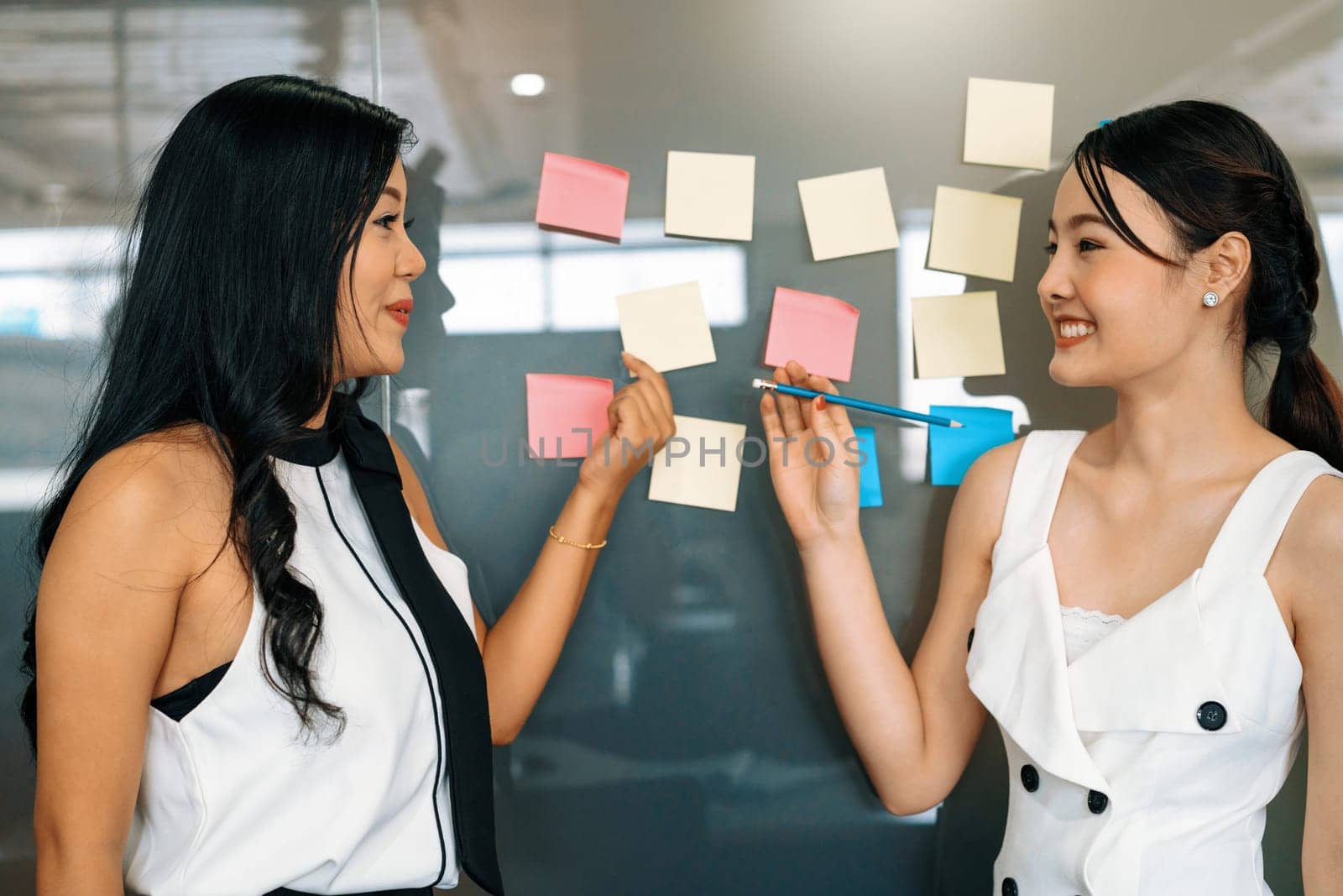 Two young beautiful Asian businesswomen talk and discuss creative post on the window. It is project reminder and thinking idea notes. uds