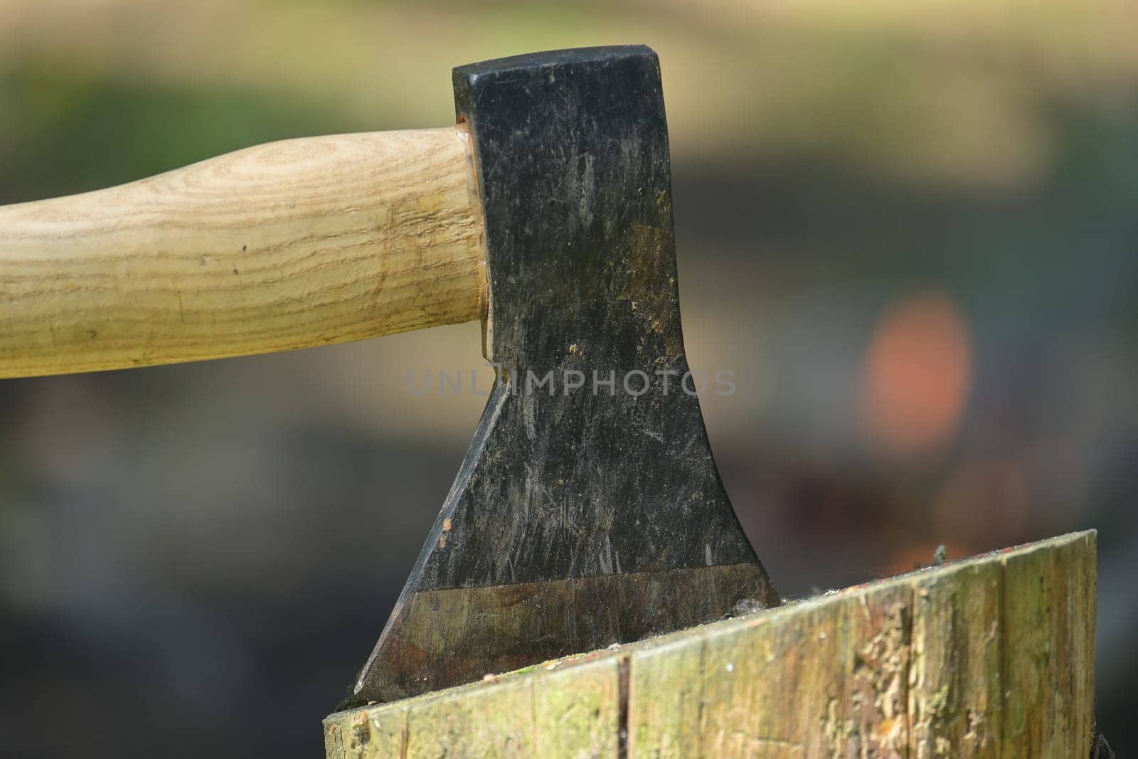Closeup view of axe with a wooden handle embedded in a log of wood, with a campfire in the background, survival or historical recreation, leisure activities and relaxation around a fire