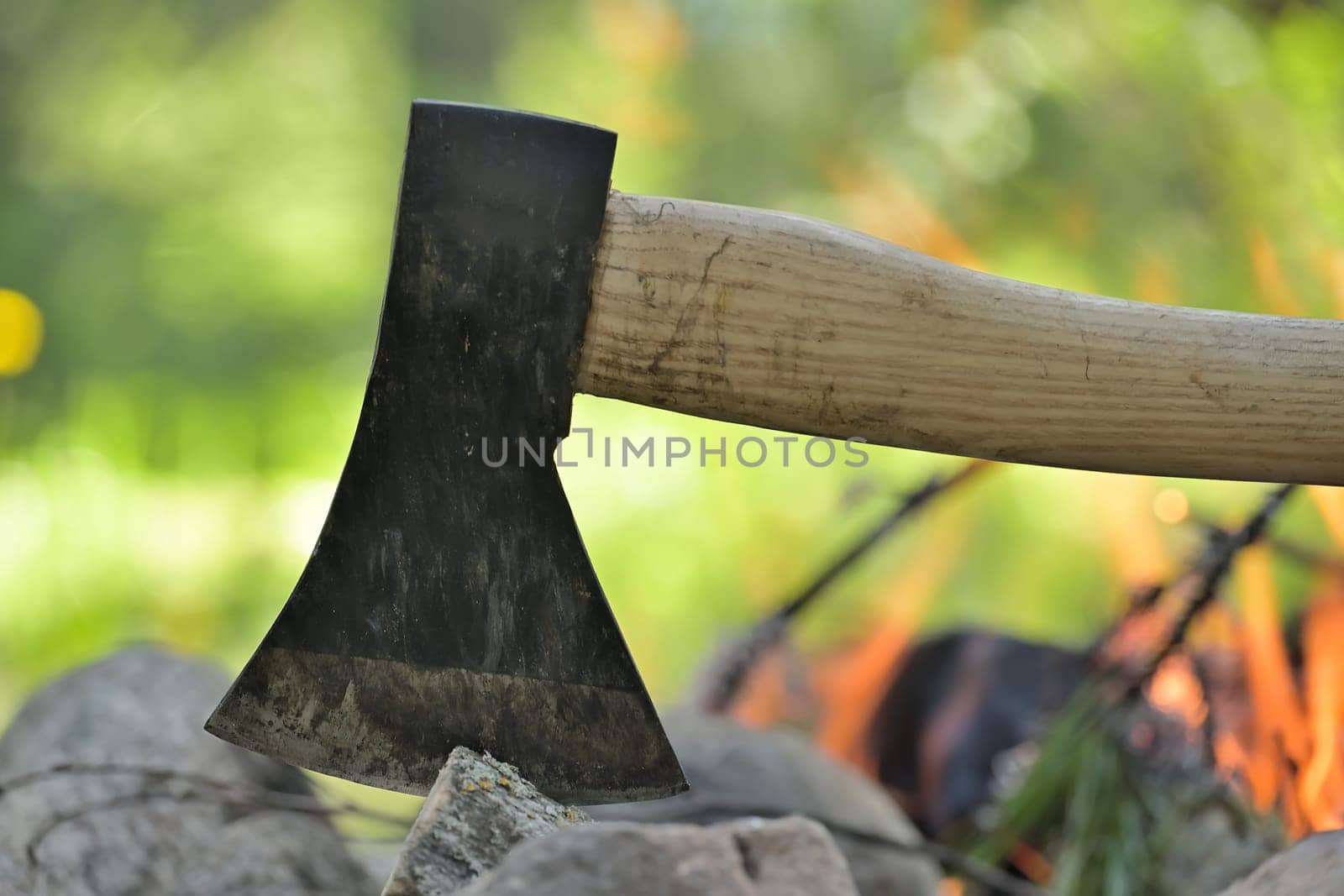 Closeup view of an axe with a wooden handle, embedded in a log, with the warm glow of a campfire in the background, survival or historical recreation, leisure activities and relaxation around a fire