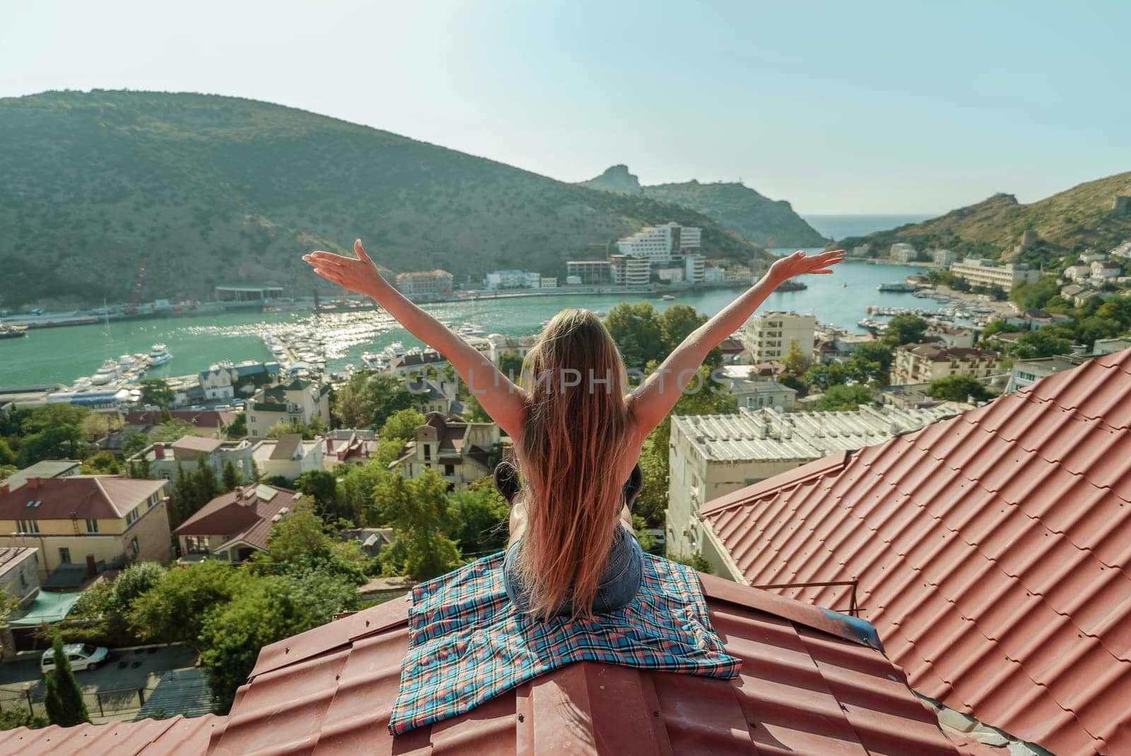 Woman sits on rooftop with outstretched arms, enjoys town view and sea mountains. Peaceful rooftop relaxation. Below her, there is a town with several boats visible in the water.